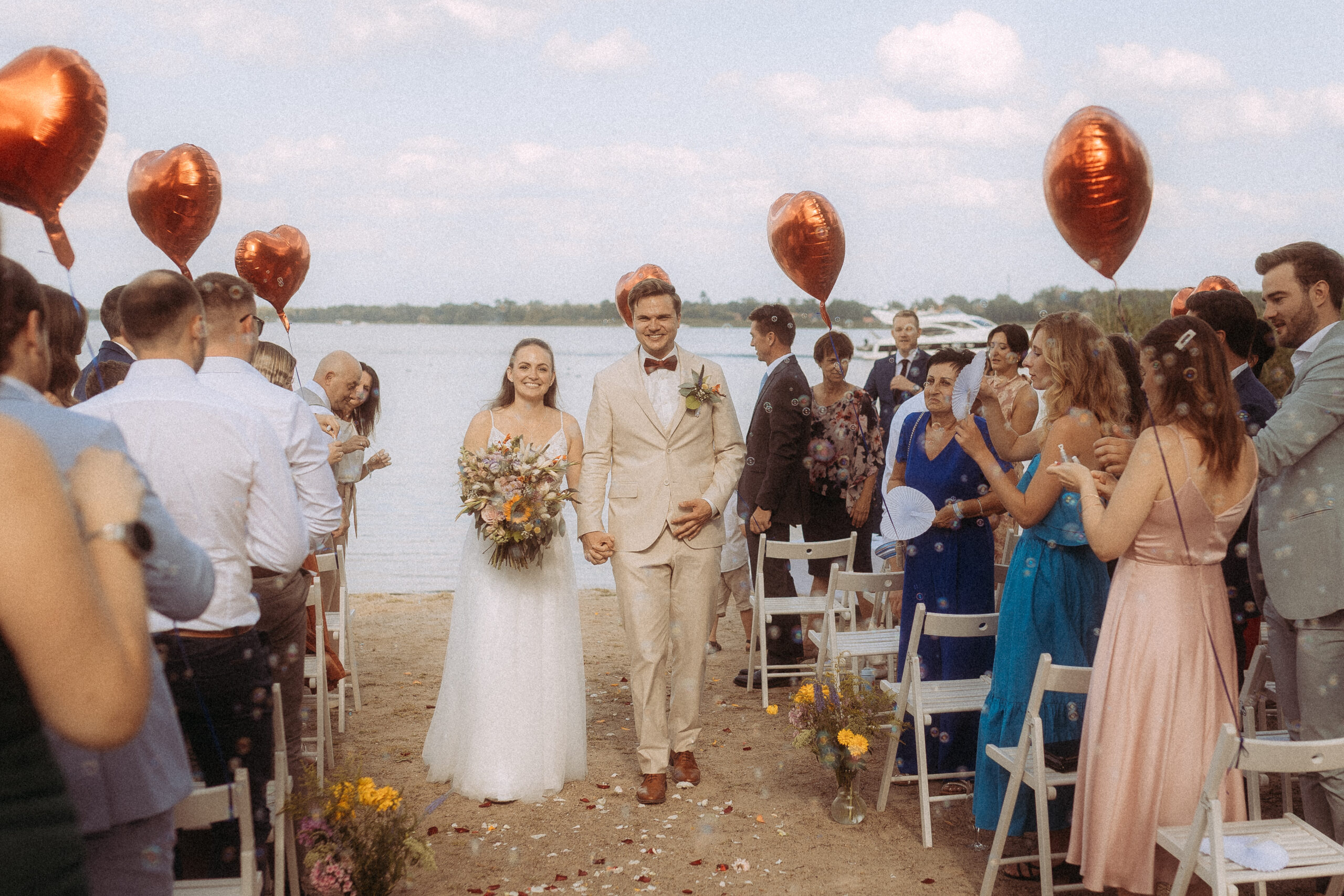 Brautpaar verlässt die Trauung am Haus Heinz am See, umgeben von Gästen und roten Herzballons