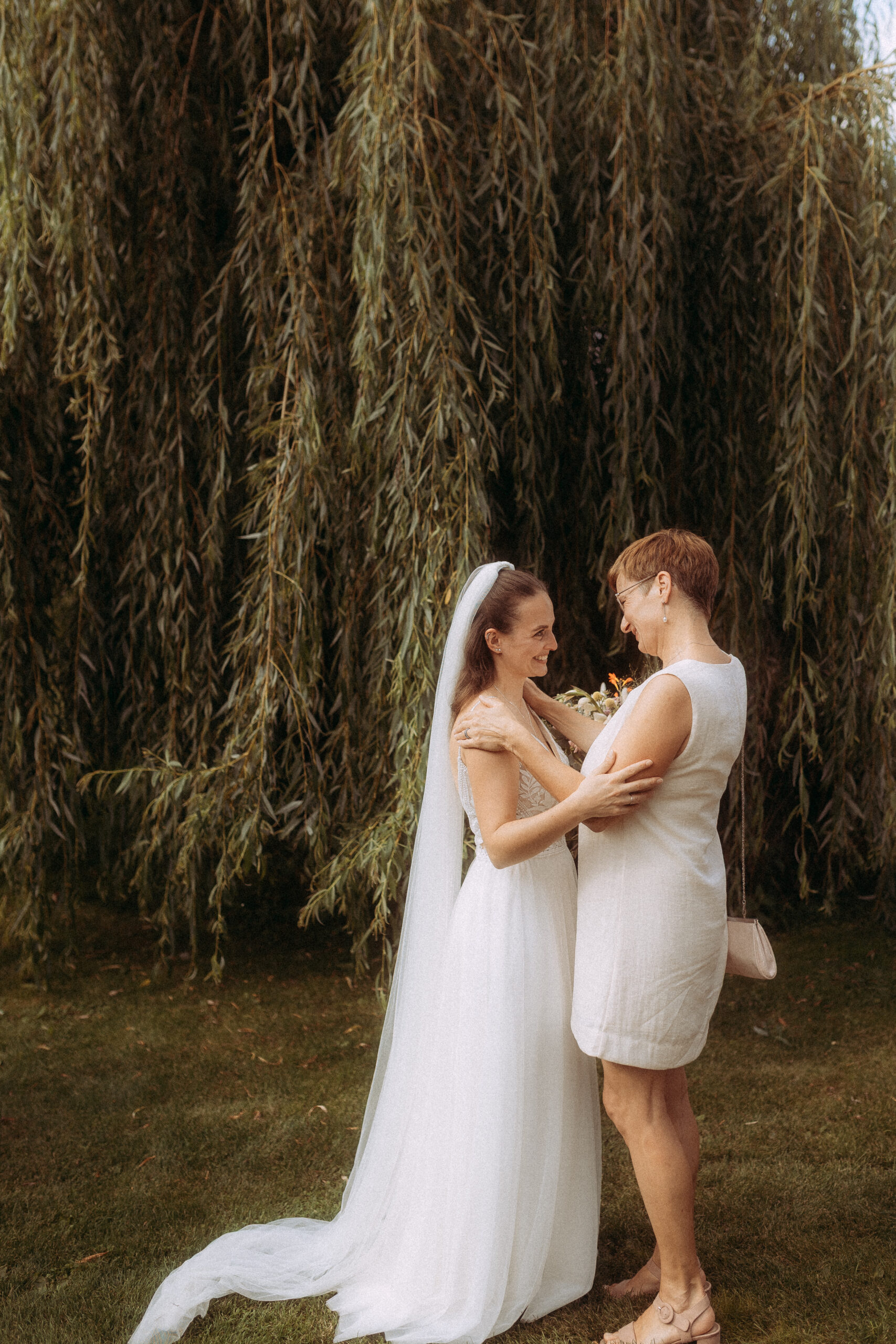 Braut und Mutter umarmen sich liebevoll vor einem Baum.