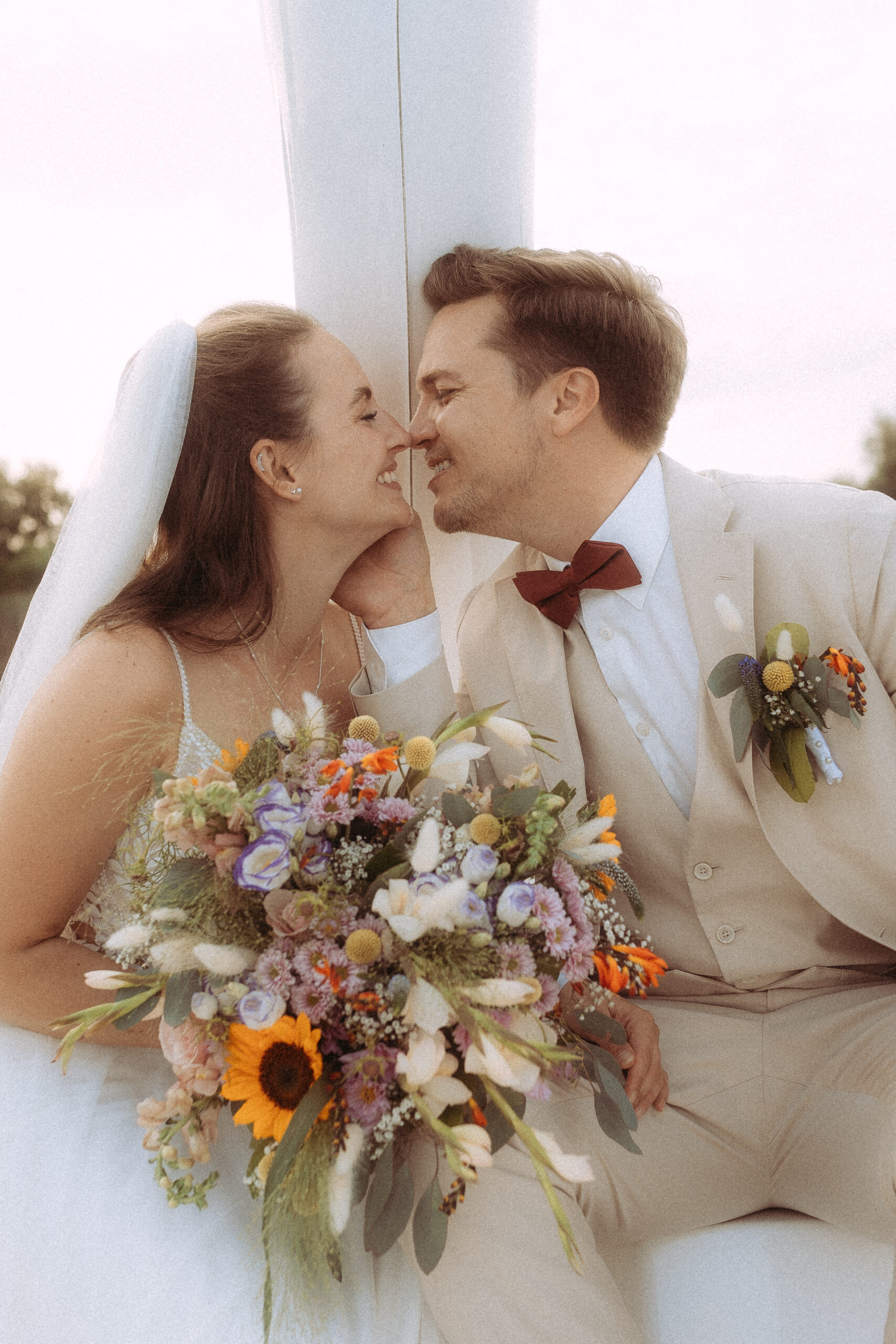 Brautpaar in beiger Hochzeitskleidung mit Blumenstrauß, lächelnd und verliebt.