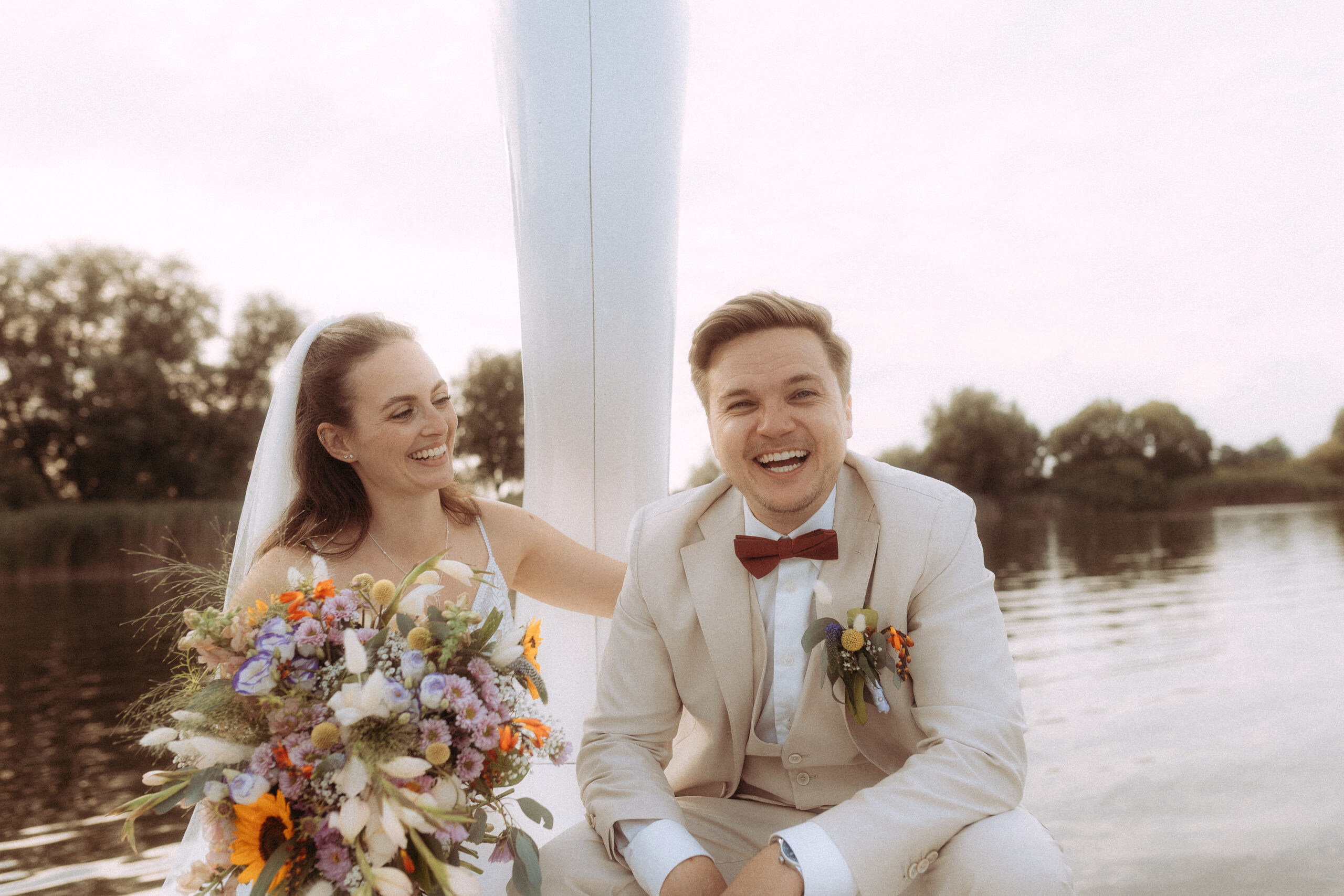 Fröhliches Brautpaar auf einem Boot, die Braut mit Blumenstrauß, der Bräutigam im hellen Anzug mit Fliege