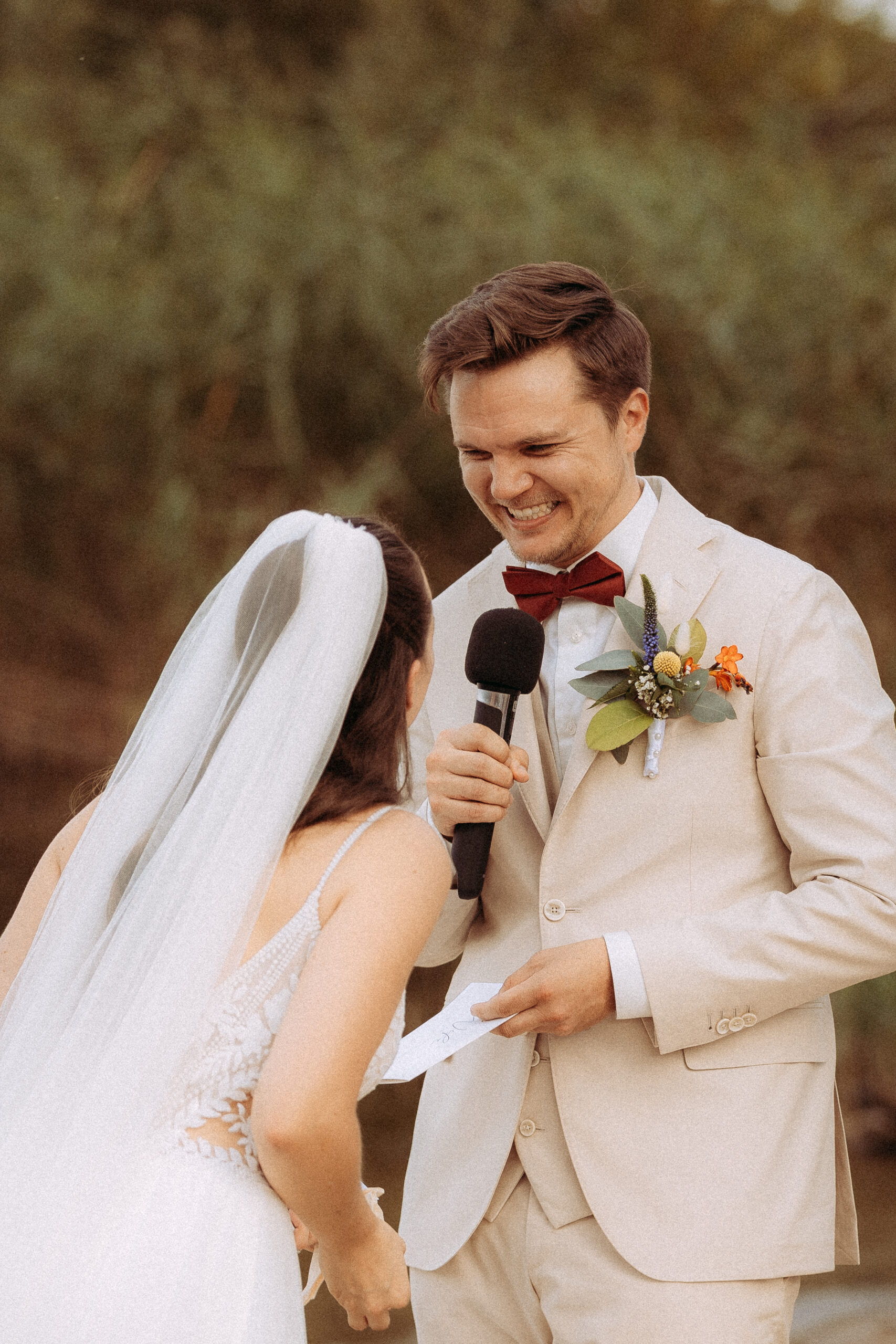 Bräutigam hält Rede mit Mikrofon für Braut bei Hochzeit im Freien.