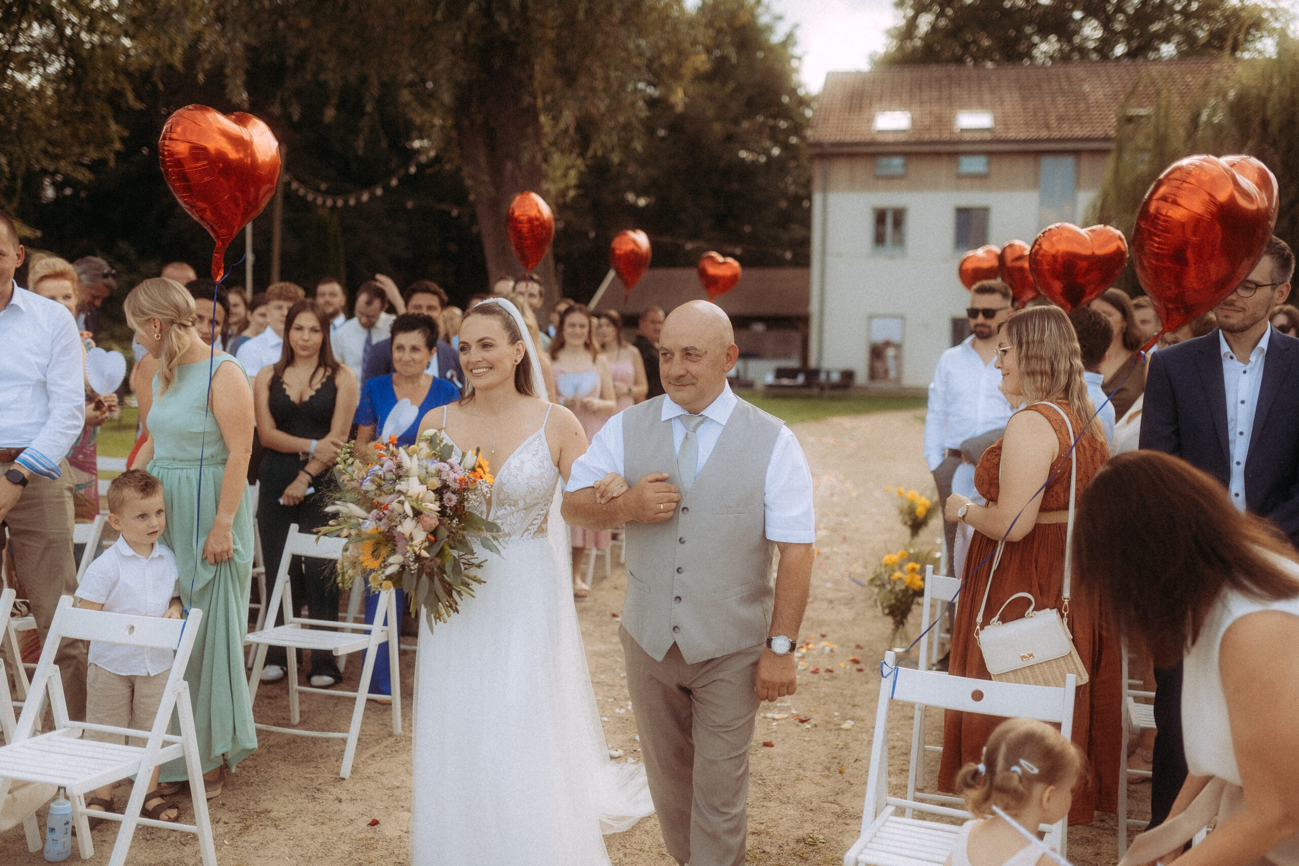 Braut mit Vater bei der Hochzeit am Haus Heinz am See, umgeben von Gästen und roten Herzballons