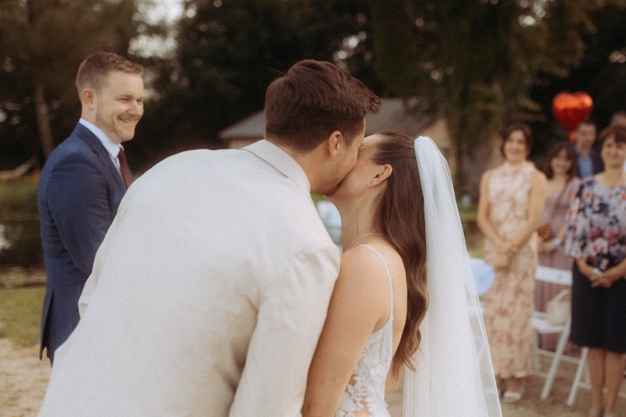 Brautpaar küsst sich bei Hochzeit im Freien, lächelnder Gast im Hintergrund.