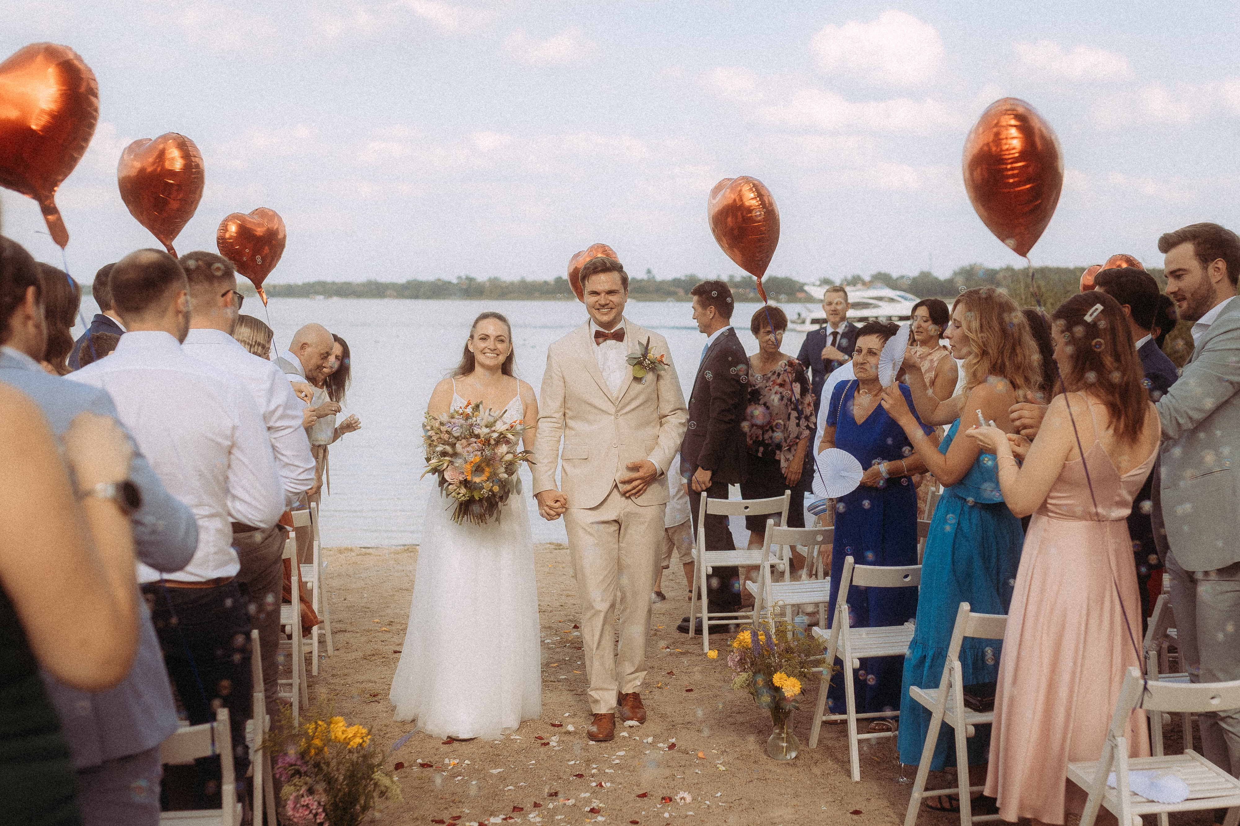 Brautpaar verlässt die Trauung am Haus Heinz am See, umgeben von Gästen und roten Herzballons