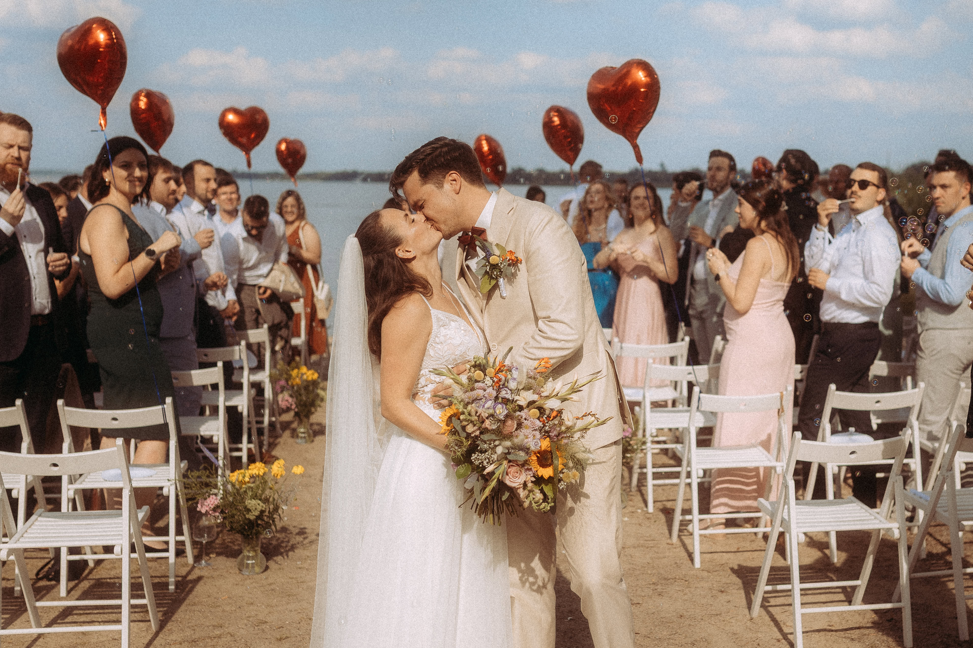 Brautpaar küsst sich am Strand, umgeben von Hochzeitsgästen und roten Herzballons.