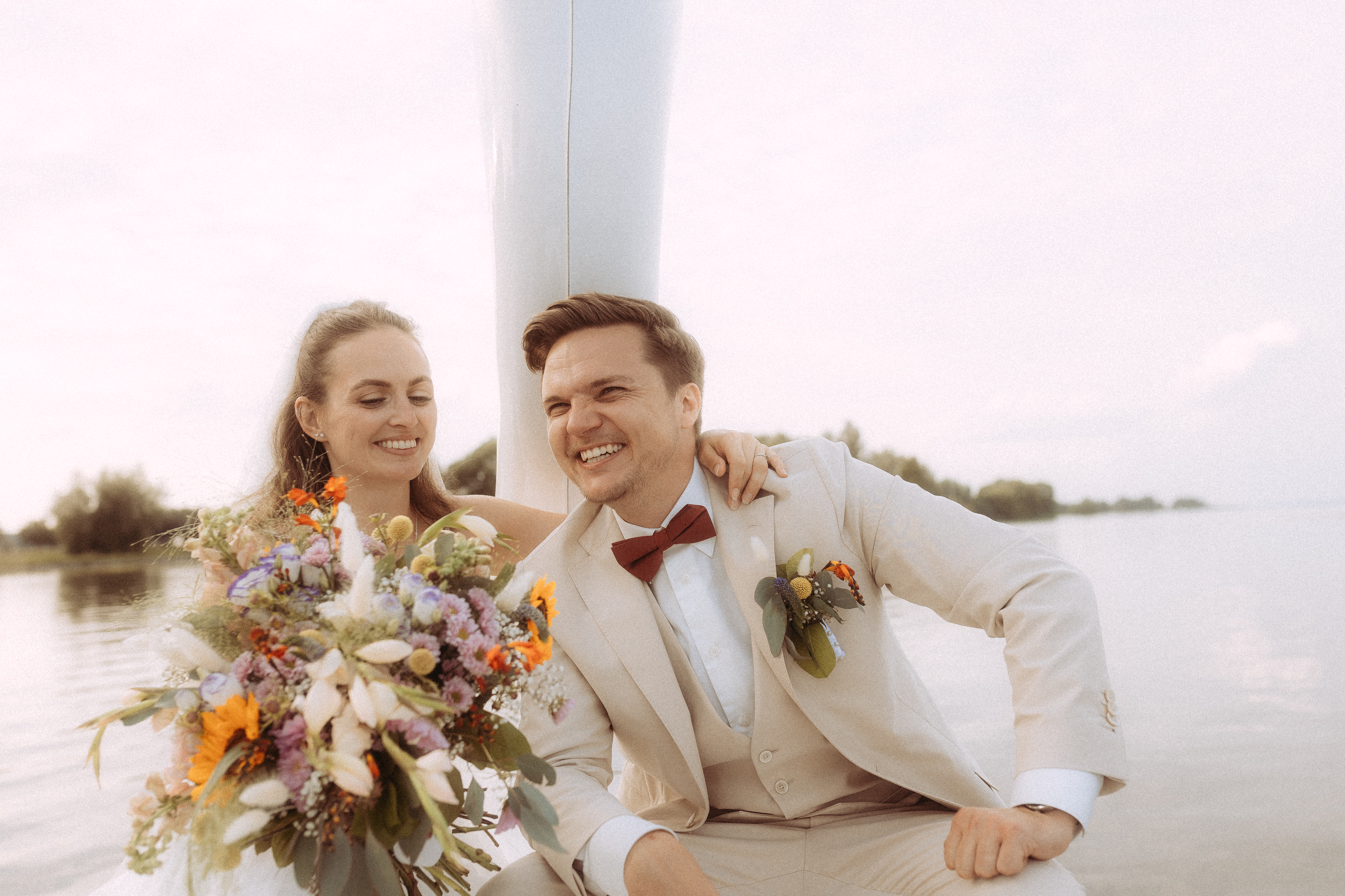 Lachendes Brautpaar mit Blumenstrauß bei Hochzeit im Freien.