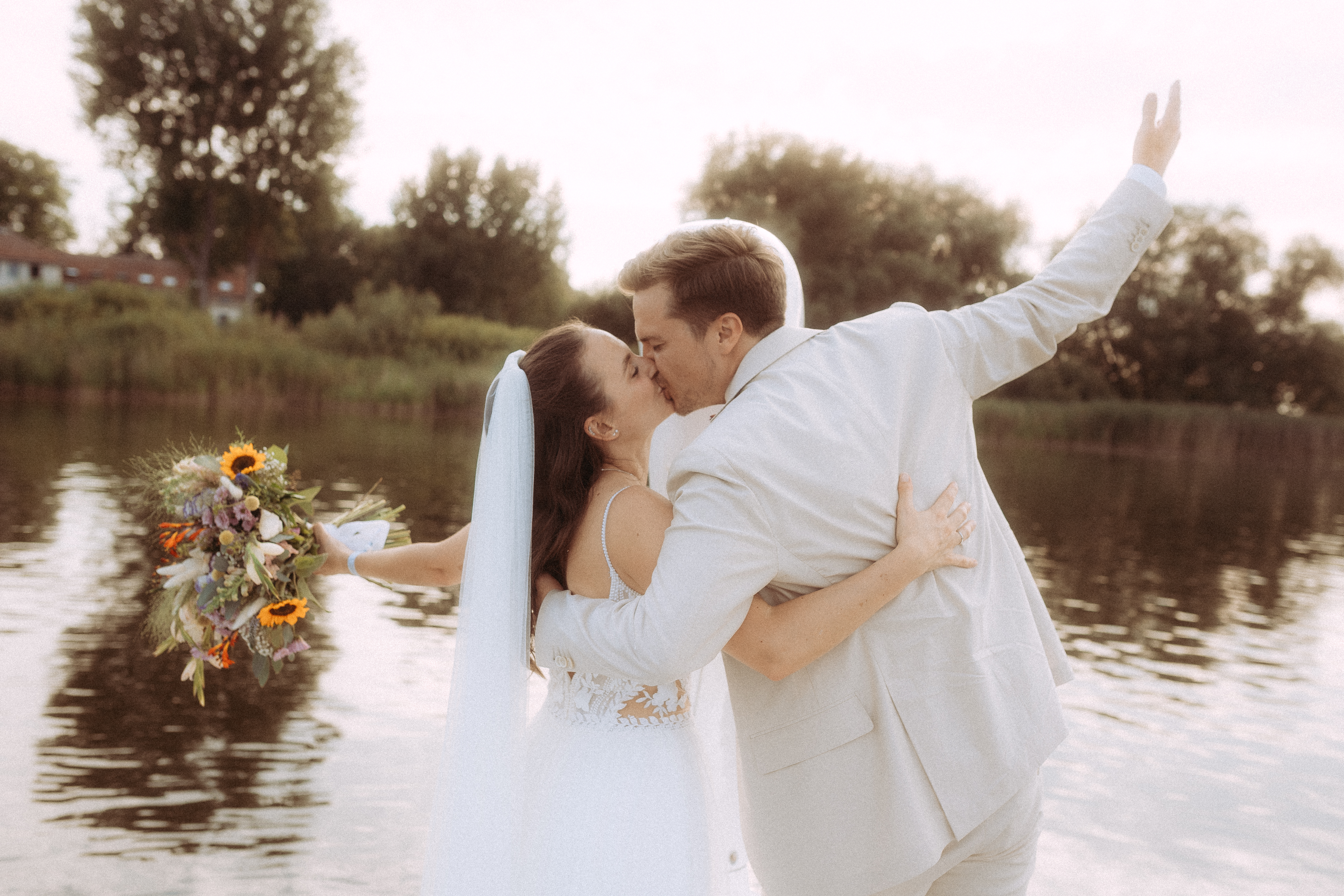 Brautpaar küsst sich am See, die Braut hält einen bunten Blumenstrauß. Hochzeit am Wasser im Freien.