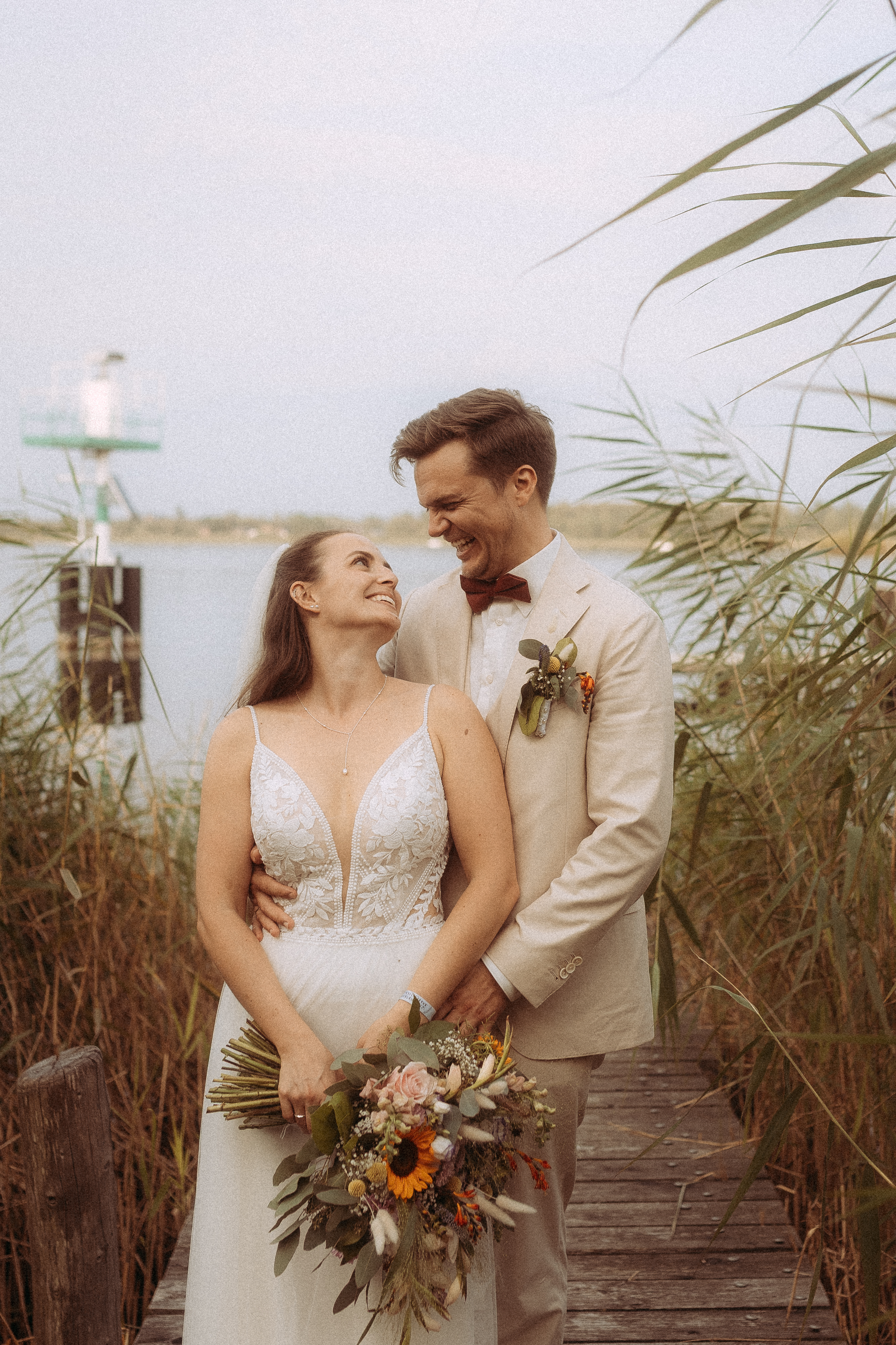Glückliches Hochzeitspaar am See, Braut mit Blumenstrauß, Bräutigam im hellen Anzug.