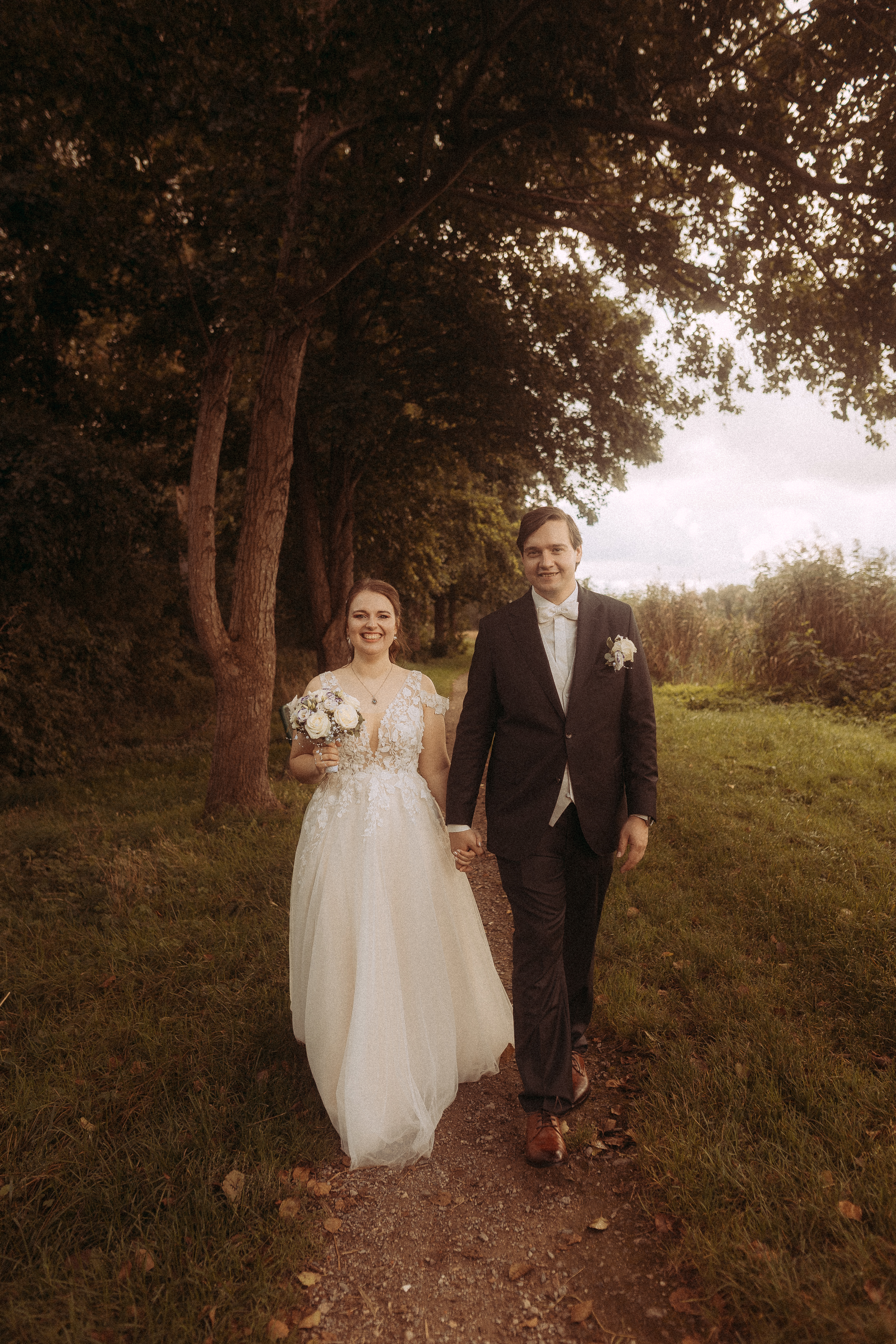 Brautpaar Hand in Hand auf Waldweg. Die Braut trägt ein weißes Kleid, der Bräutigam einen dunklen Anzug.