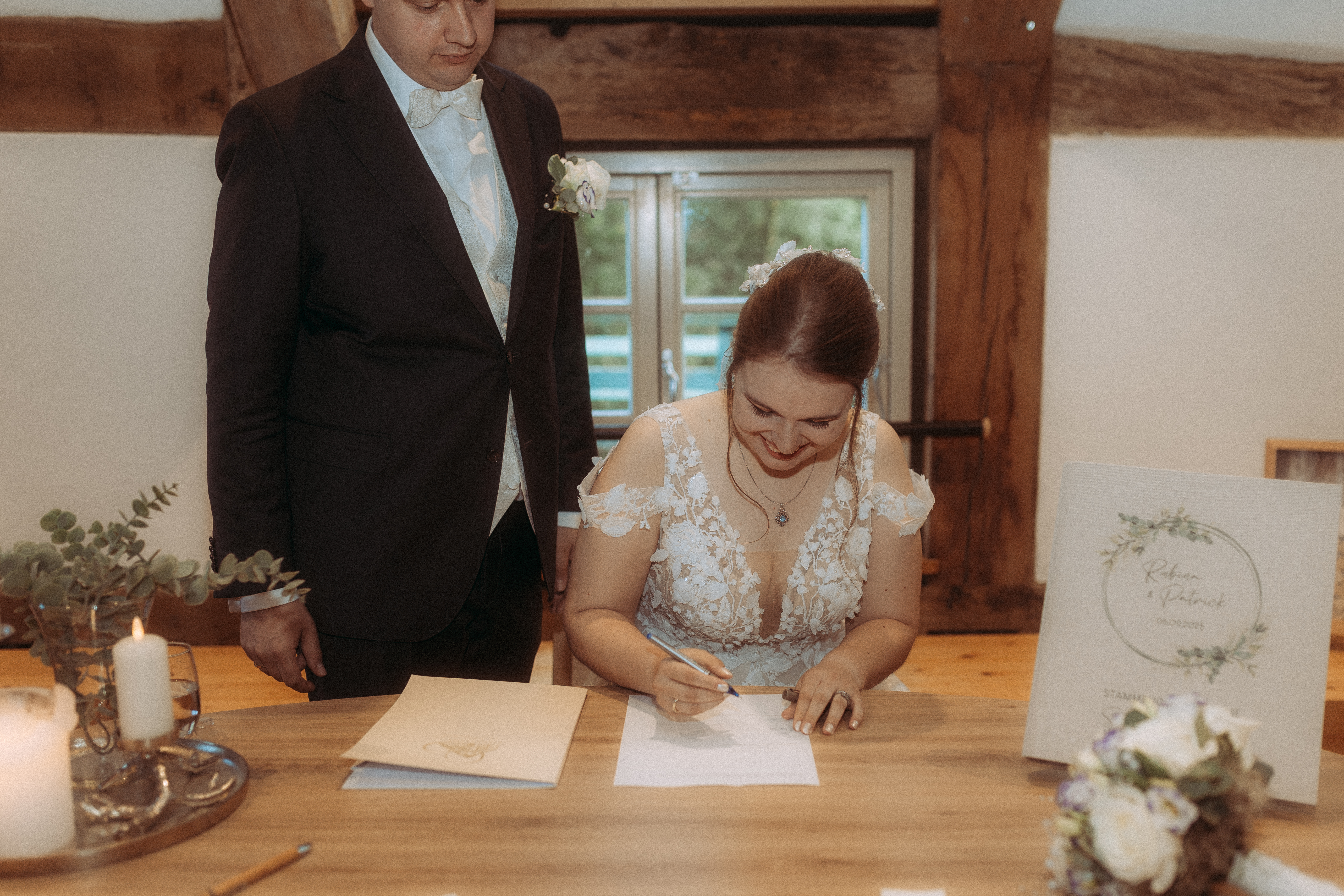 Braut unterschreibt Dokument bei der Hochzeit am Haus Heinz am See, Bräutigam steht daneben.