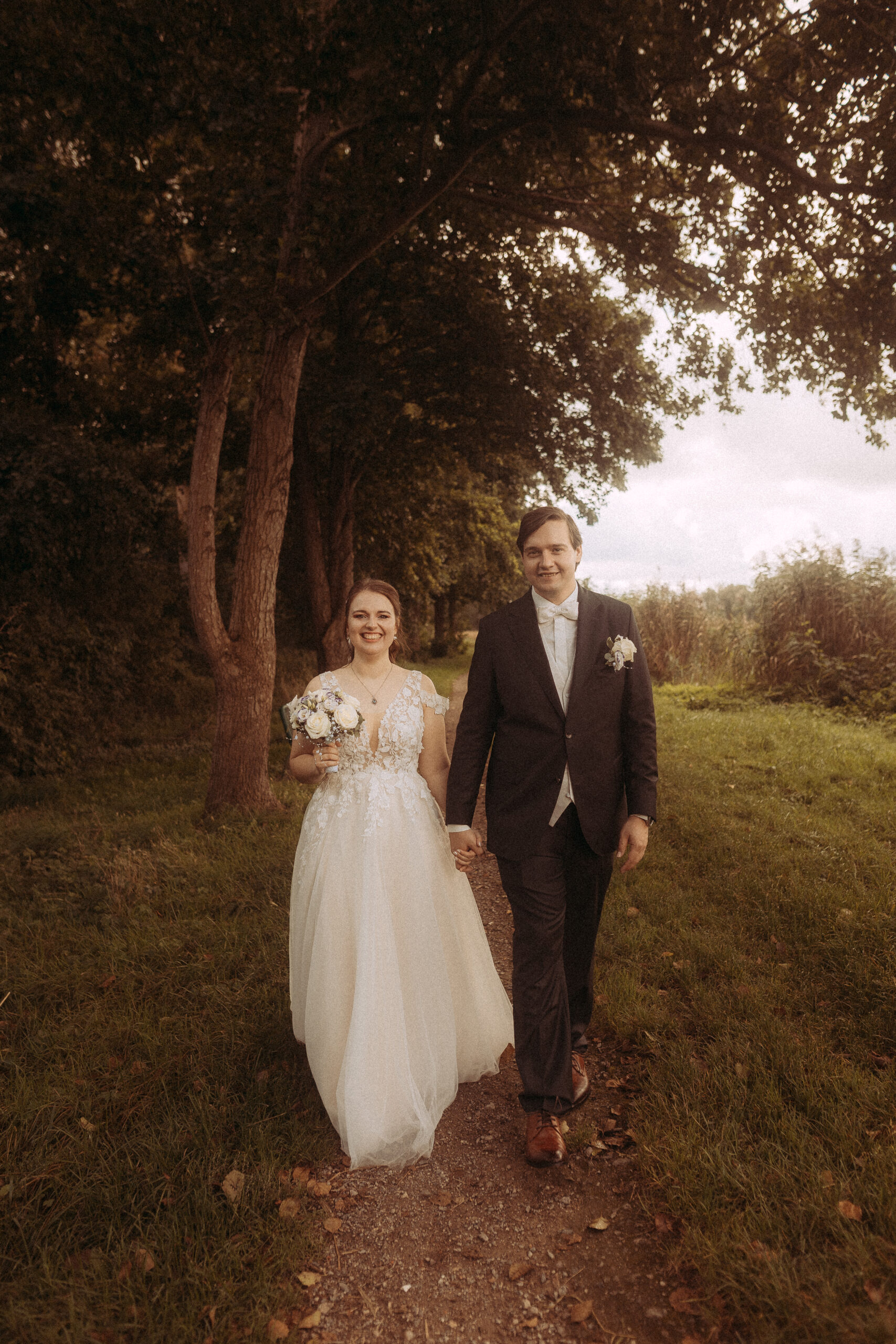 Brautpaar Hand in Hand auf Waldweg. Die Braut trägt ein weißes Kleid, der Bräutigam einen dunklen Anzug.
