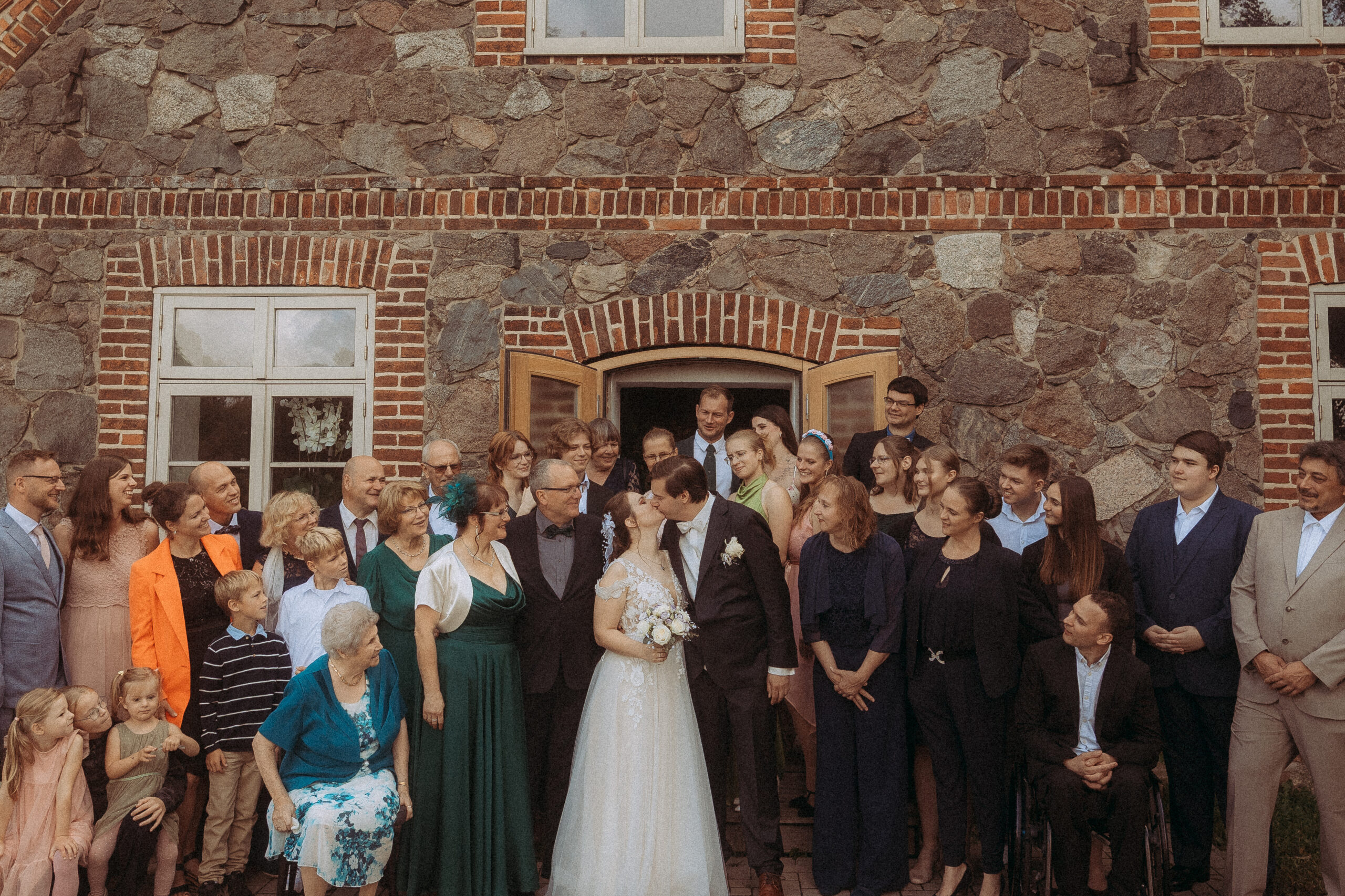 Hochzeitsgesellschaft küsst sich vor Haus Heinz am See, rustikale Steinmauer im Hintergrund