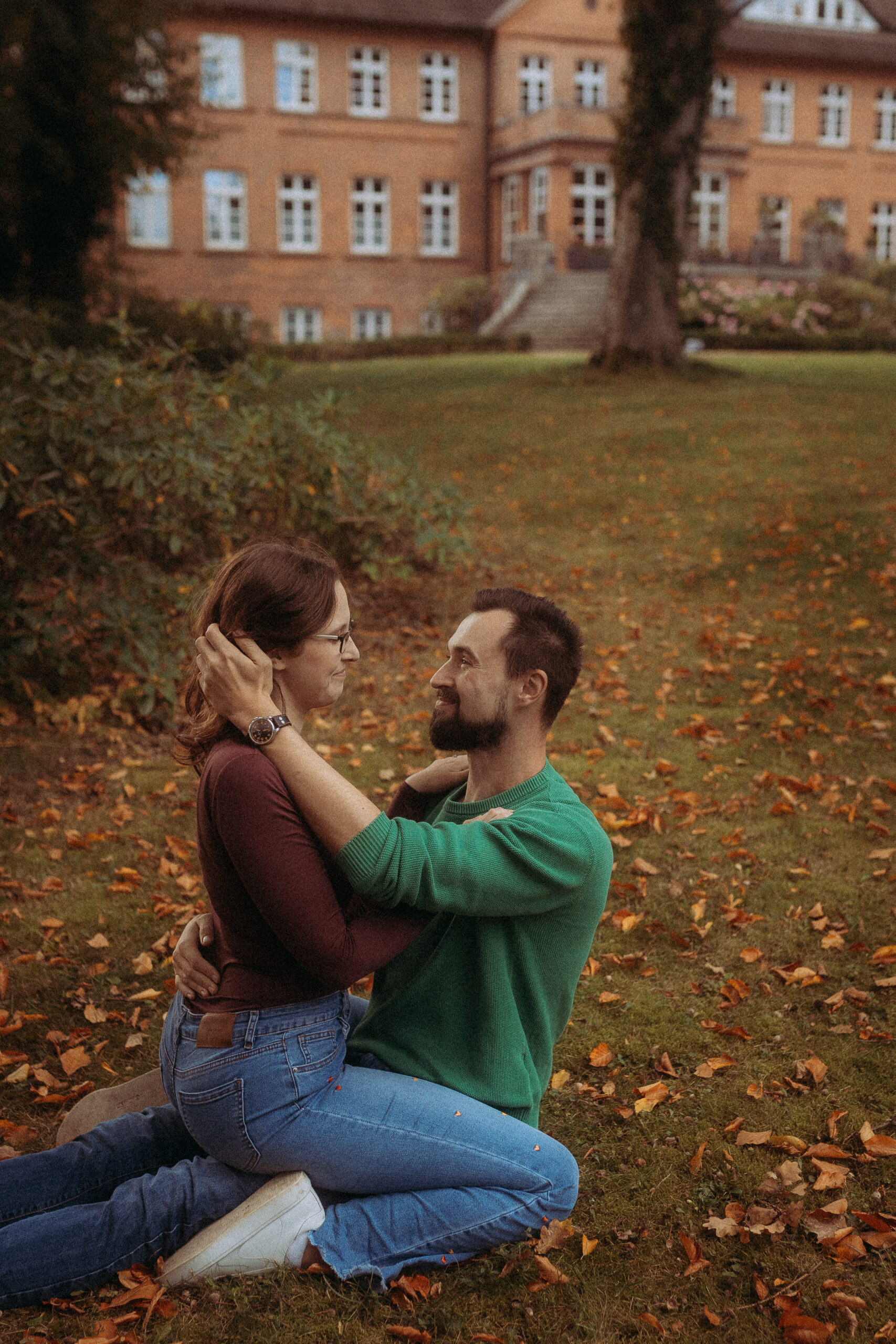 Verliebtes Paar umarmt sich auf einer Wiese vor einem Backsteingebäude. Herbstlaub liegt herum.