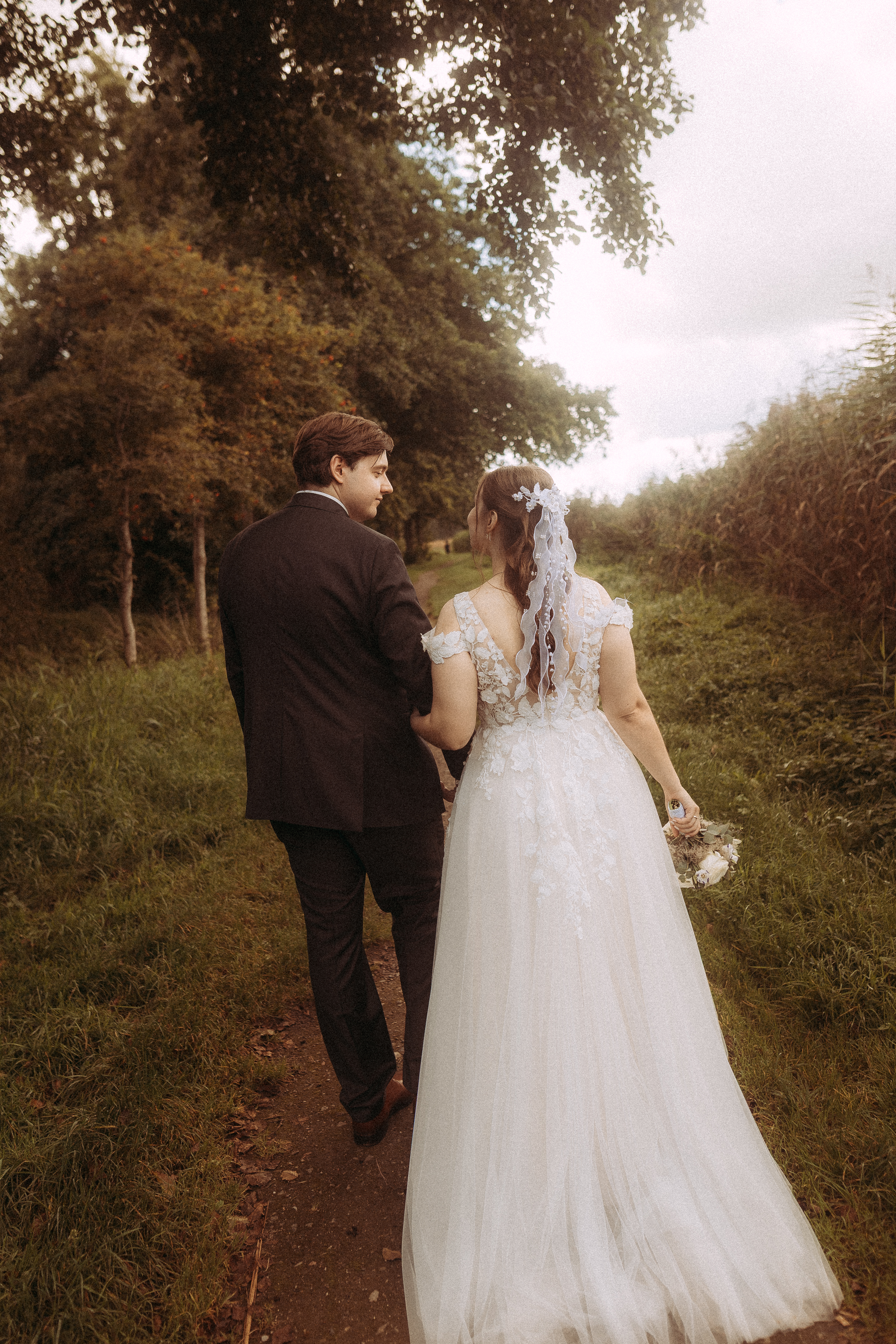 Brautpaar spaziert Händchen haltend auf einem Feldweg, Hochzeit im Grünen
