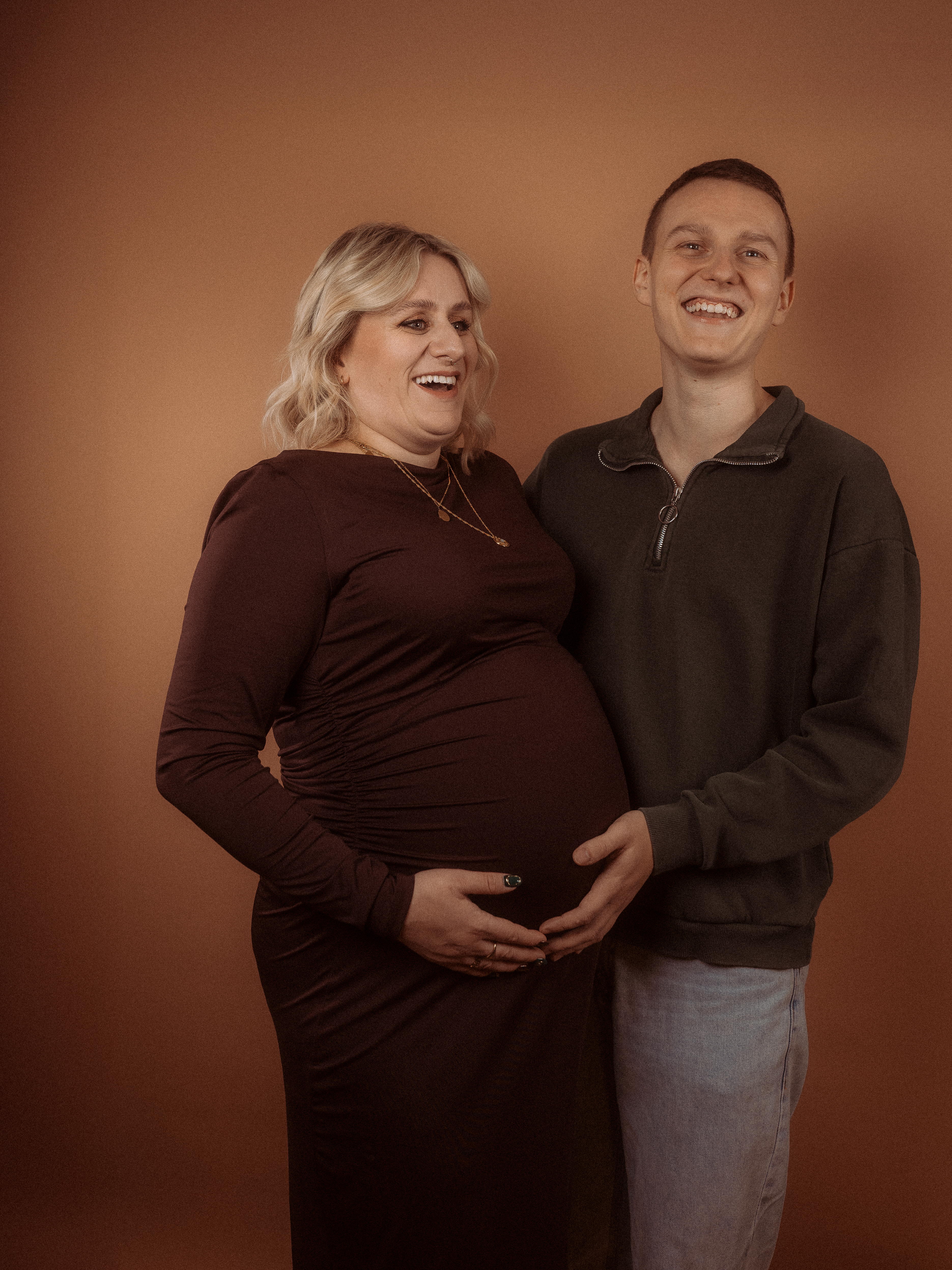 Glückliches Paar erwartet Baby in Parchim. Frau mit Babybauch im braunen Kleid, Mann hält Bauch.