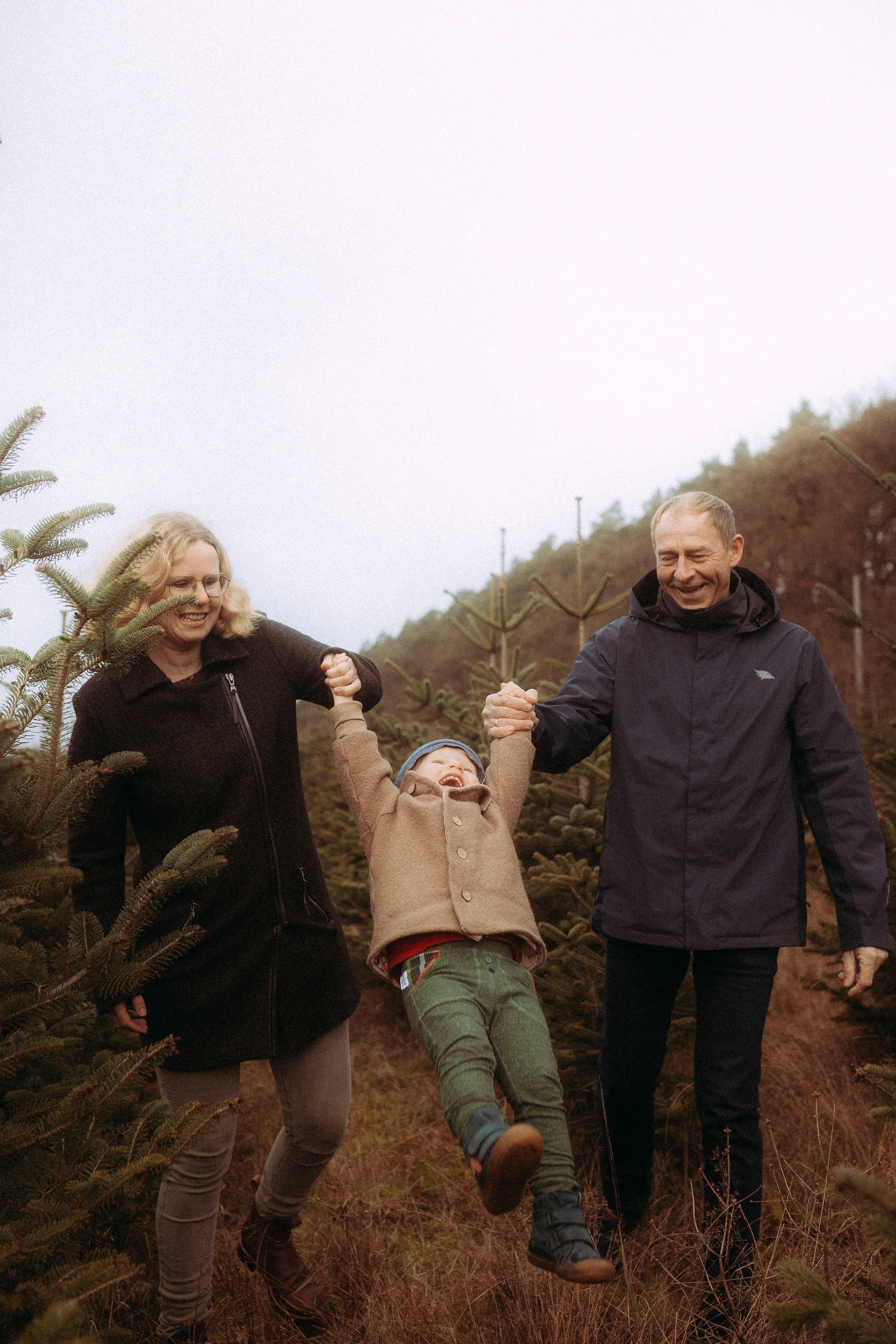 Familie hat Spaß beim Baumkauf: Eltern schwingen Kind auf Weihnachtsbaumplantage.