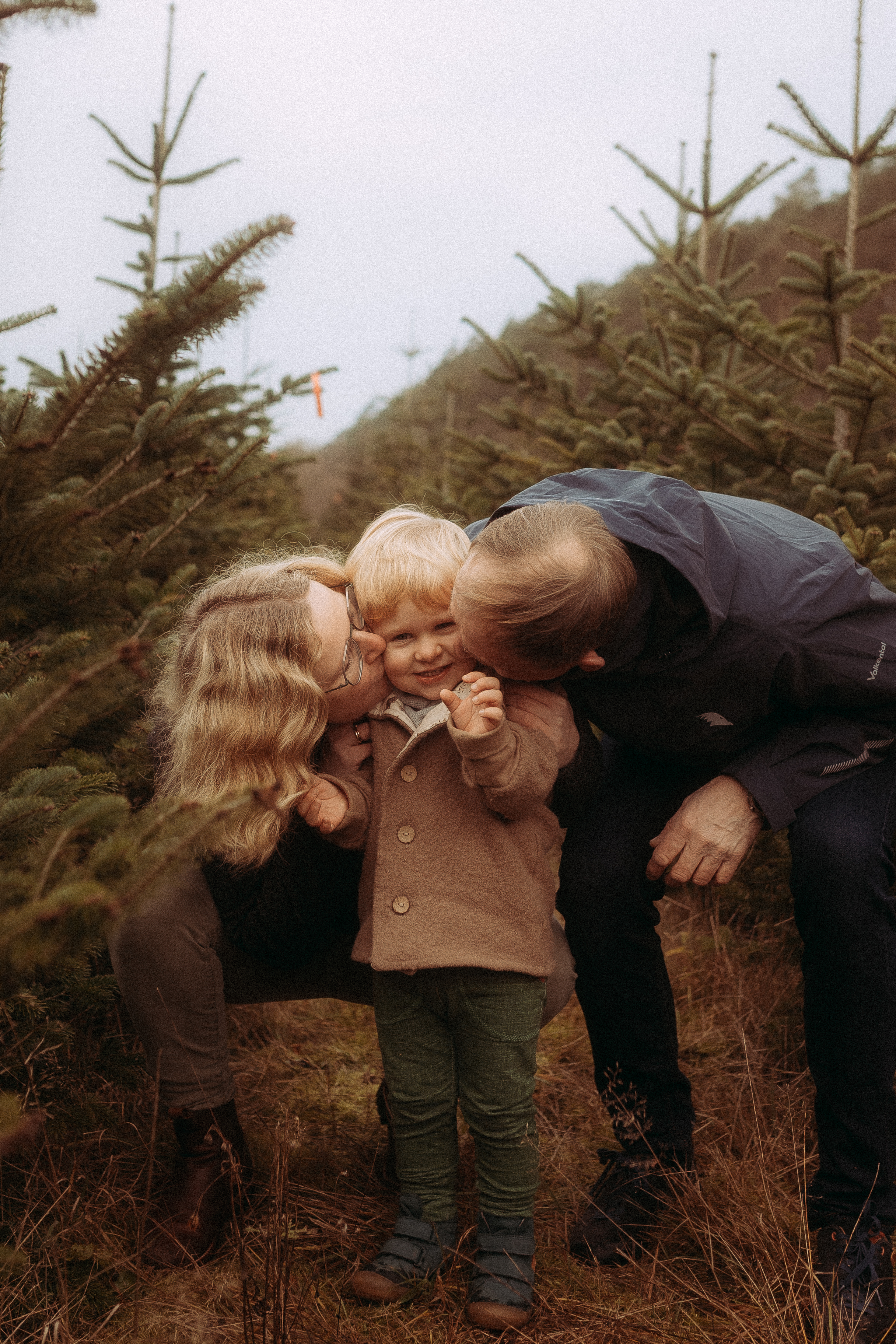 Familie umarmt Kind auf Weihnachtsbaumfarm. Eltern küssen ihren Sohn zwischen Tannenbäumen.