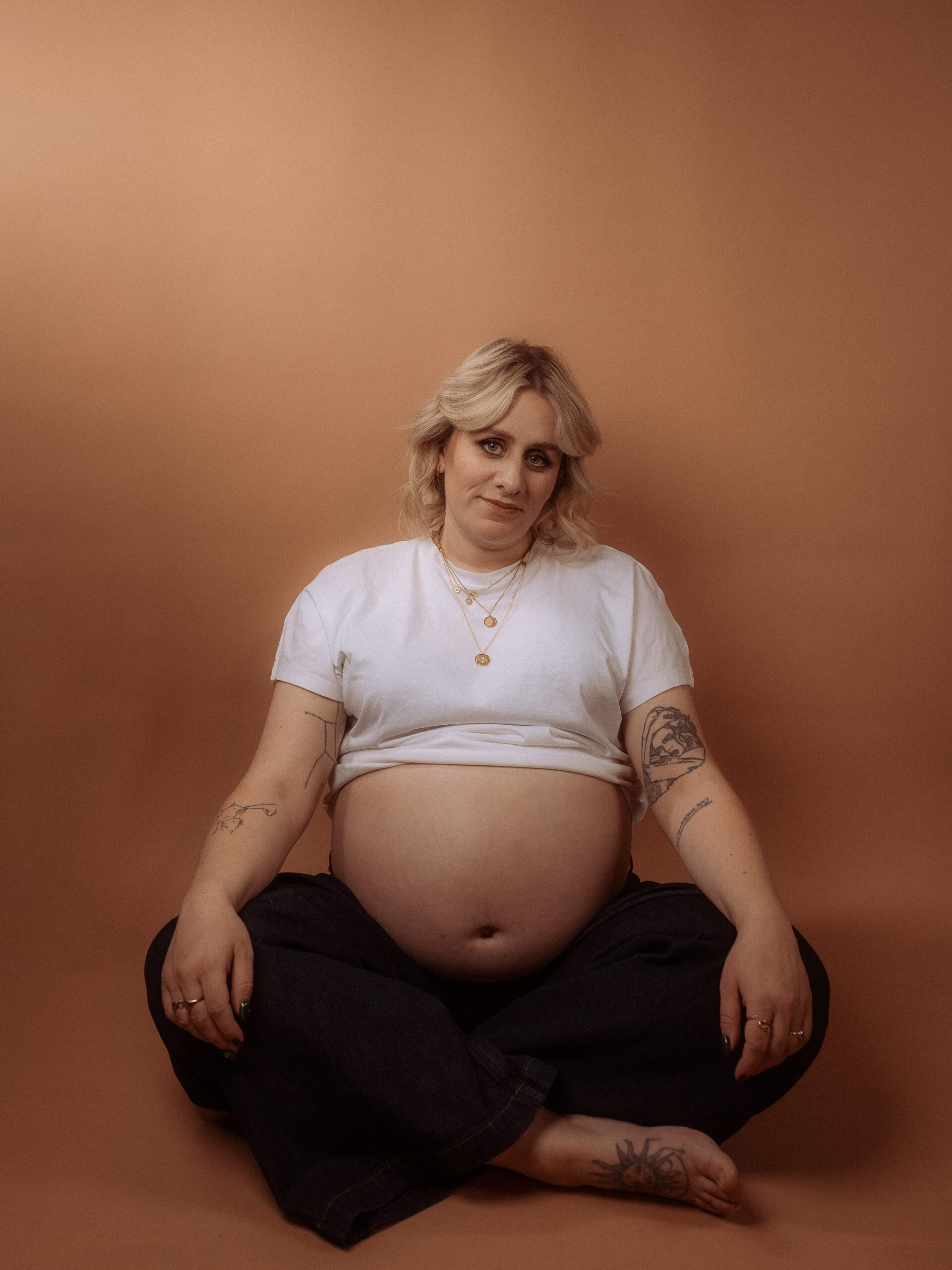 Schwangere Frau sitzt im Schneidersitz vor braunem Hintergrund. Babybauchshooting in Parchim.