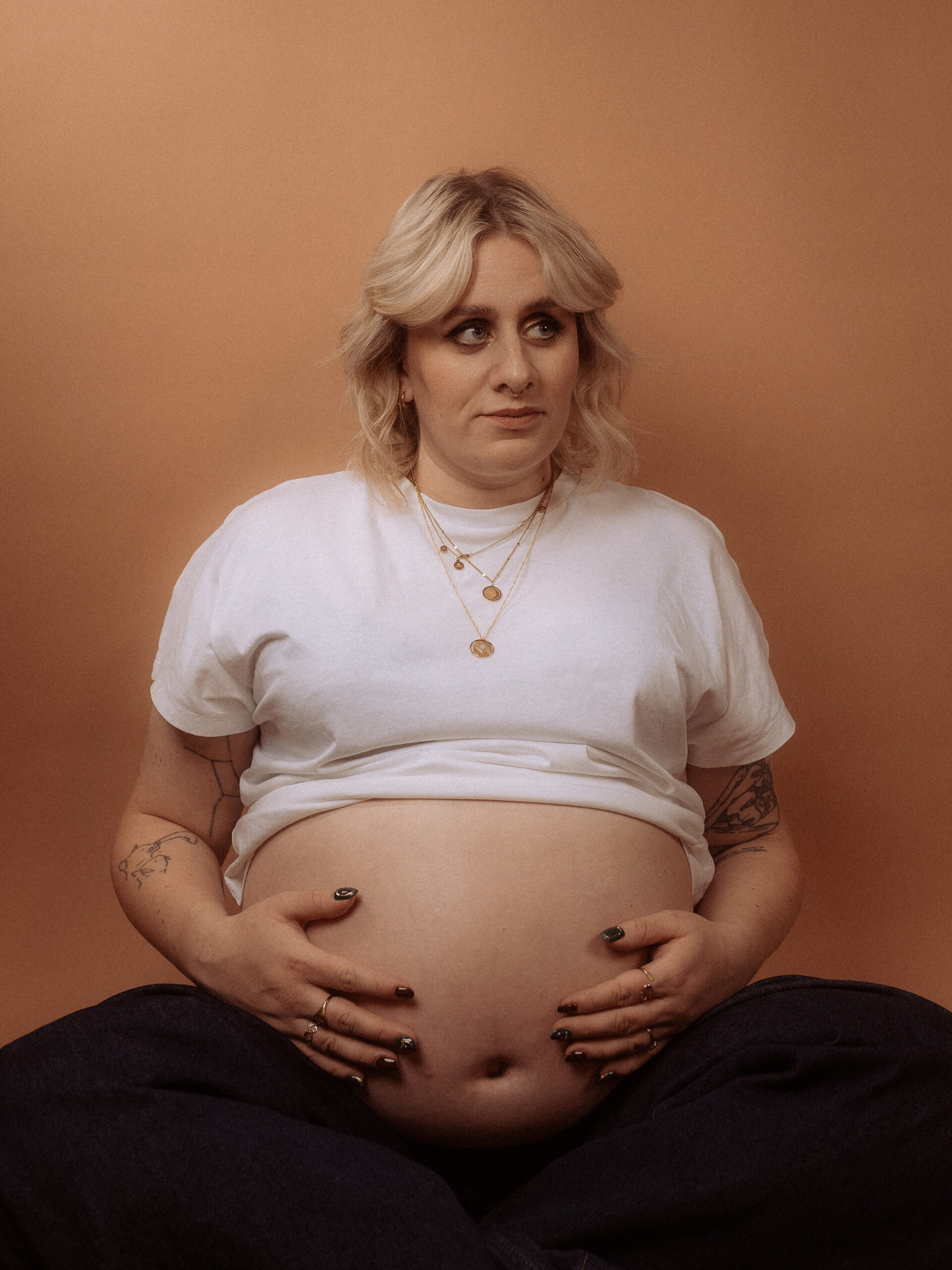 Schwangere Frau hält ihren Bauch, trägt Schmuck und ein weißes T-Shirt.