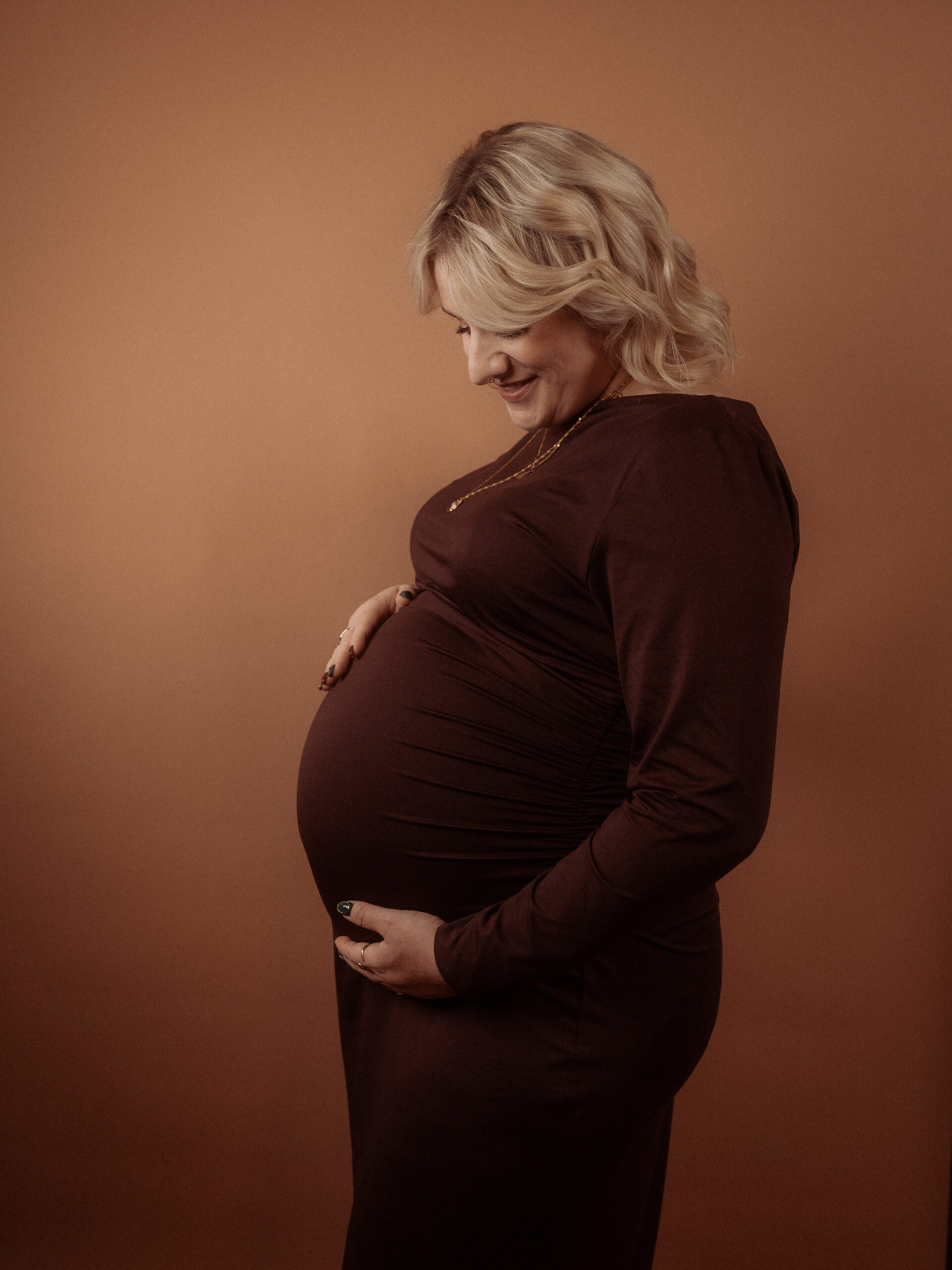 Schwangere Frau im braunen Kleid hält ihren Bauch. Familienfotografie.