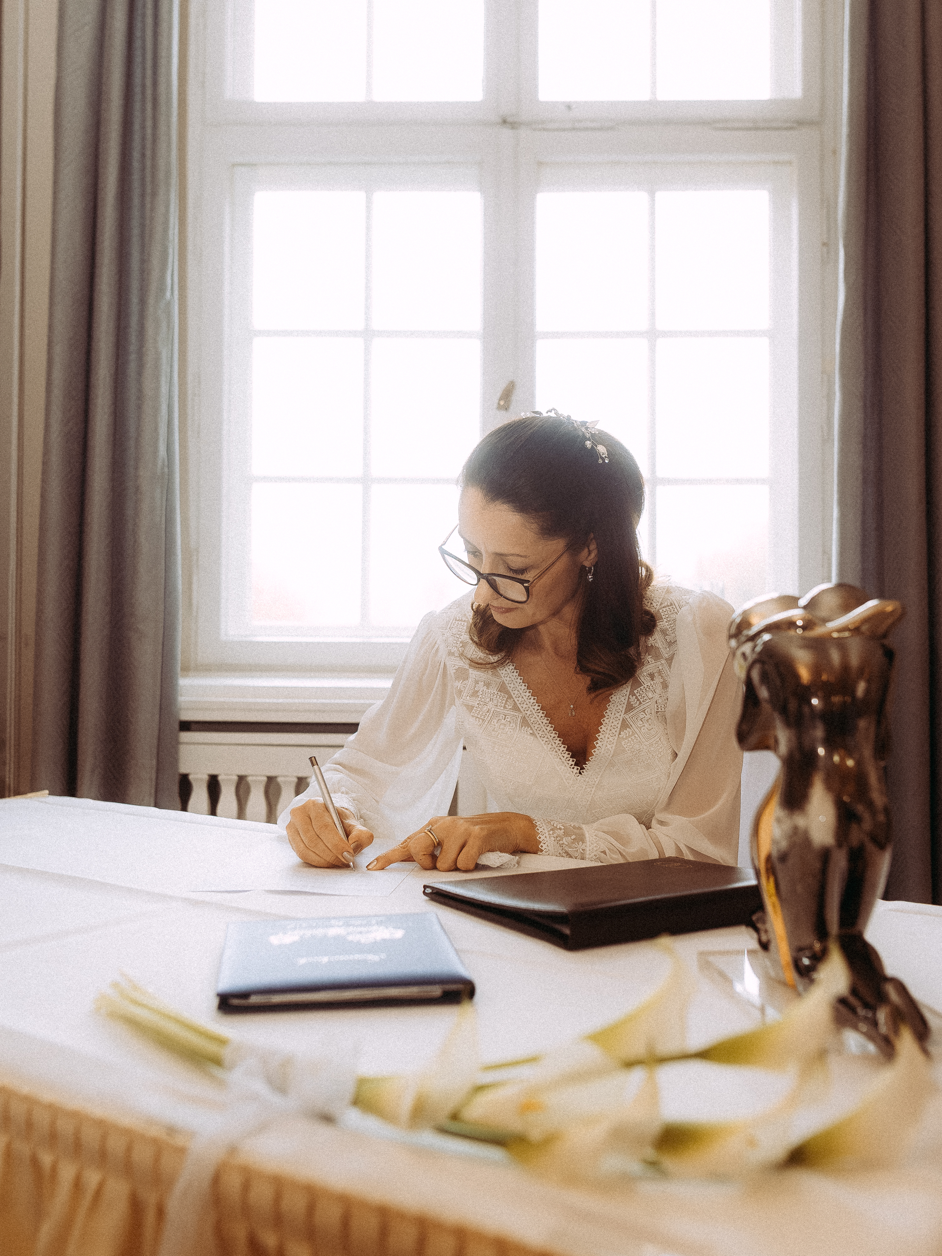 Frau unterschreibt Dokument bei einer Hochzeit. Elegante Braut mit Brille vor Fenster.