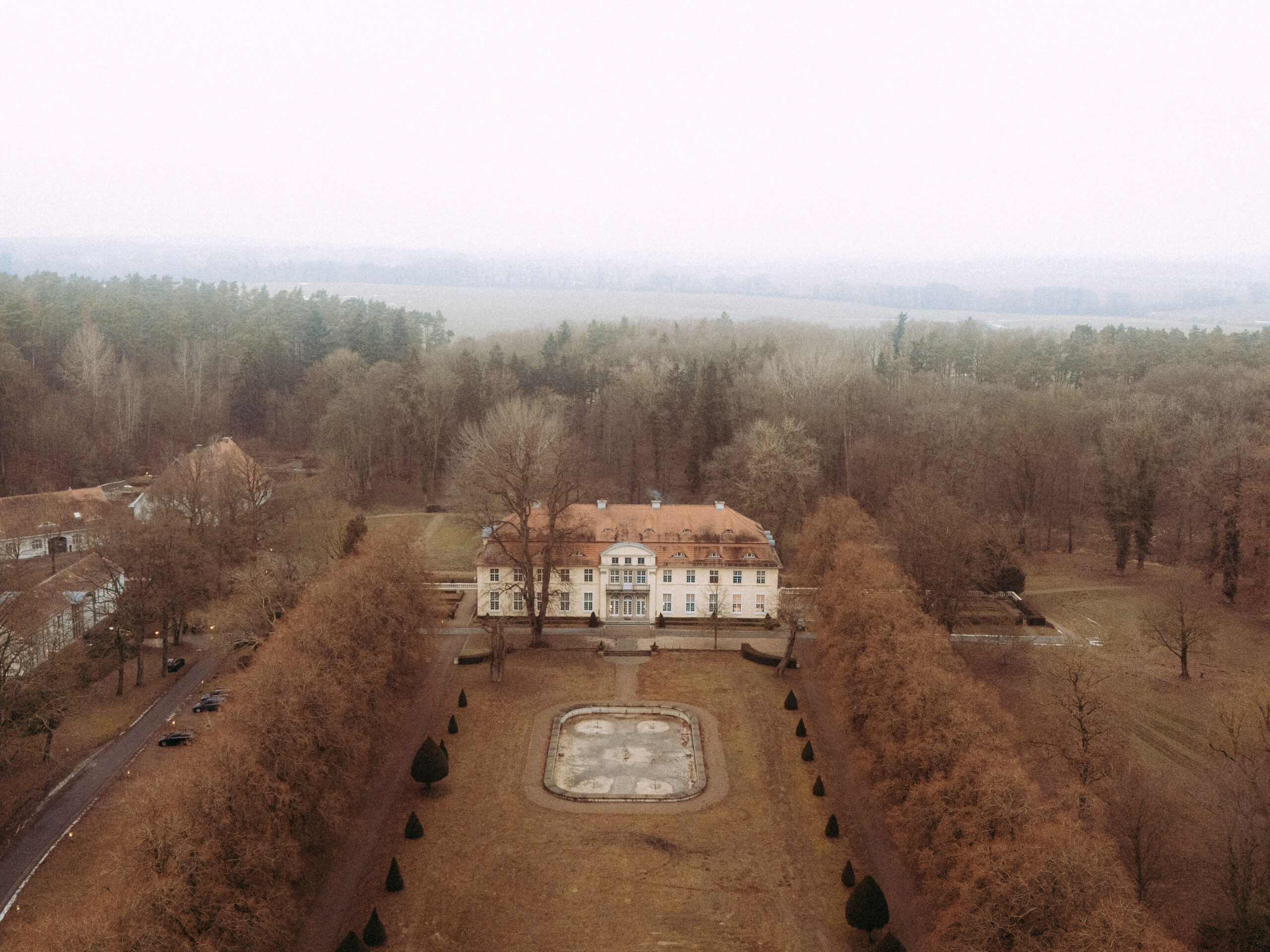 Luftaufnahme eines Schlosses mit Parkanlage und Allee an einem nebligen Tag
