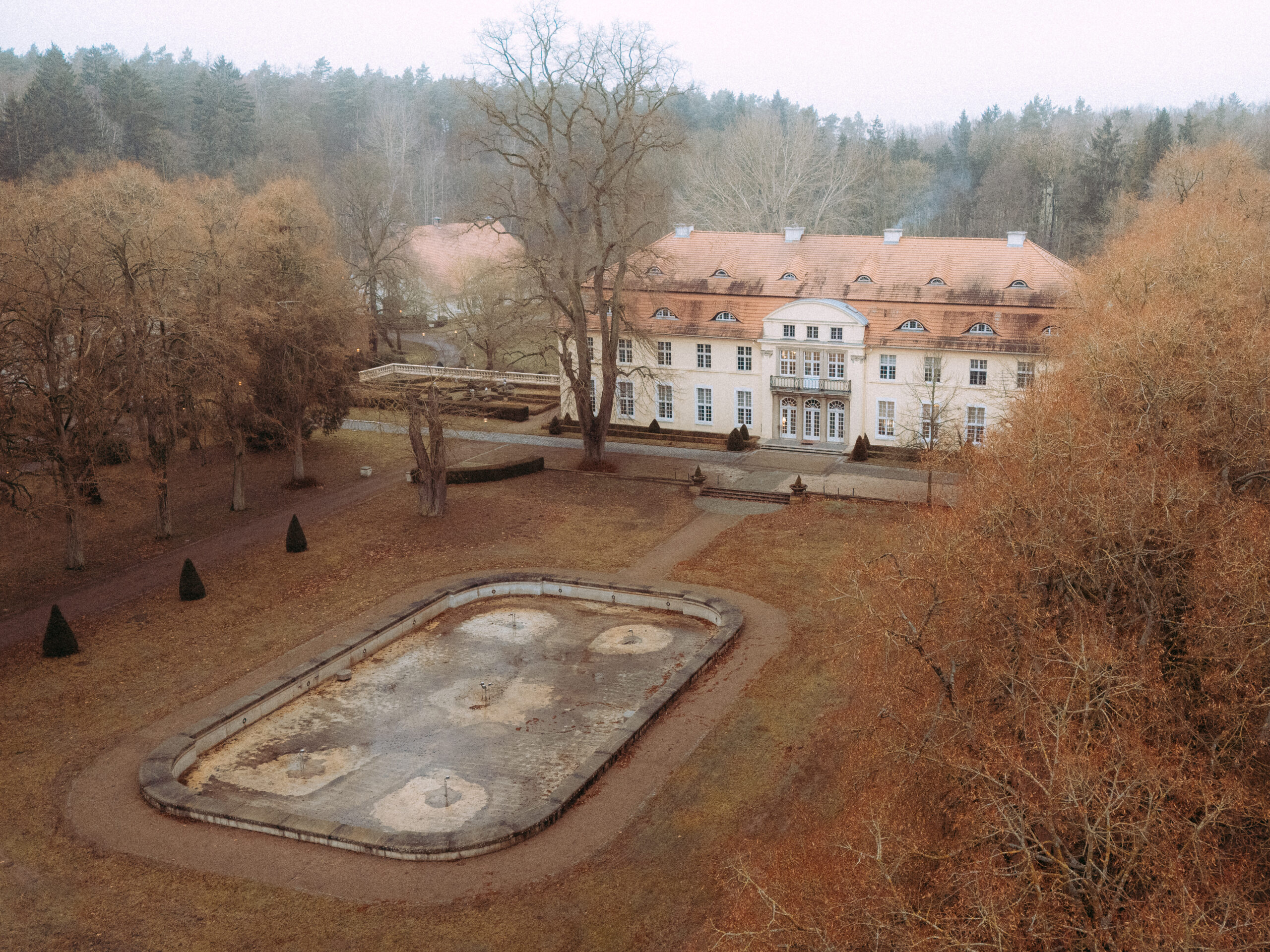 Großes Herrenhaus mit Gartenanlage und leerem Brunnenbecken im Vordergrund.