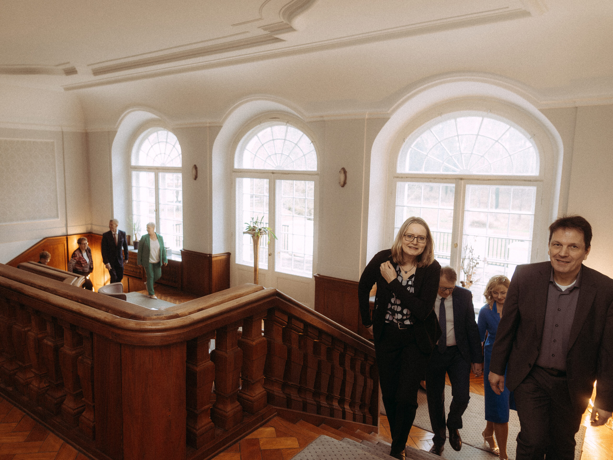 Personen steigen eine Holztreppe in einem hellen Gebäude mit großen Fenstern hinauf.