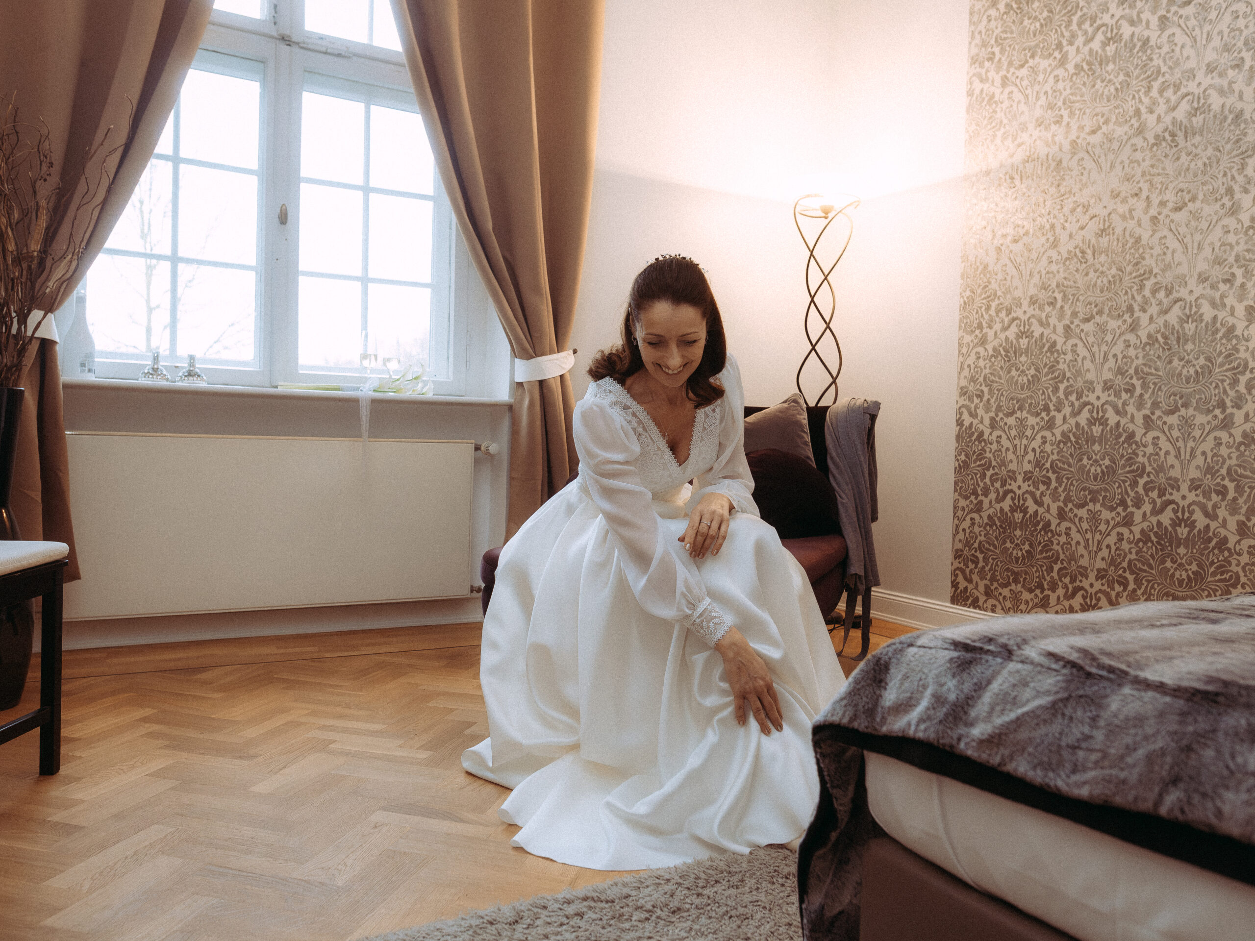 Braut im weißen Brautkleid sitzt lächelnd in einem hellen Zimmer. Hochzeit Vorbereitung.