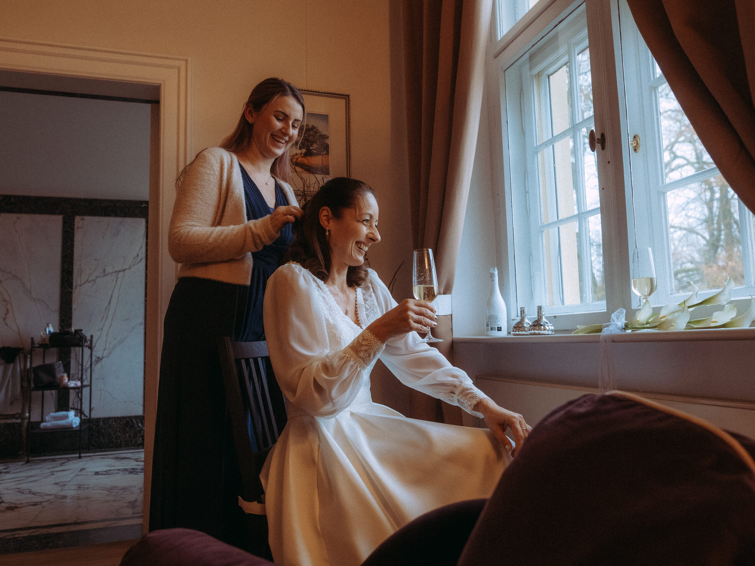 Woman in white dress getting ready, holding champagne, with friend assisting