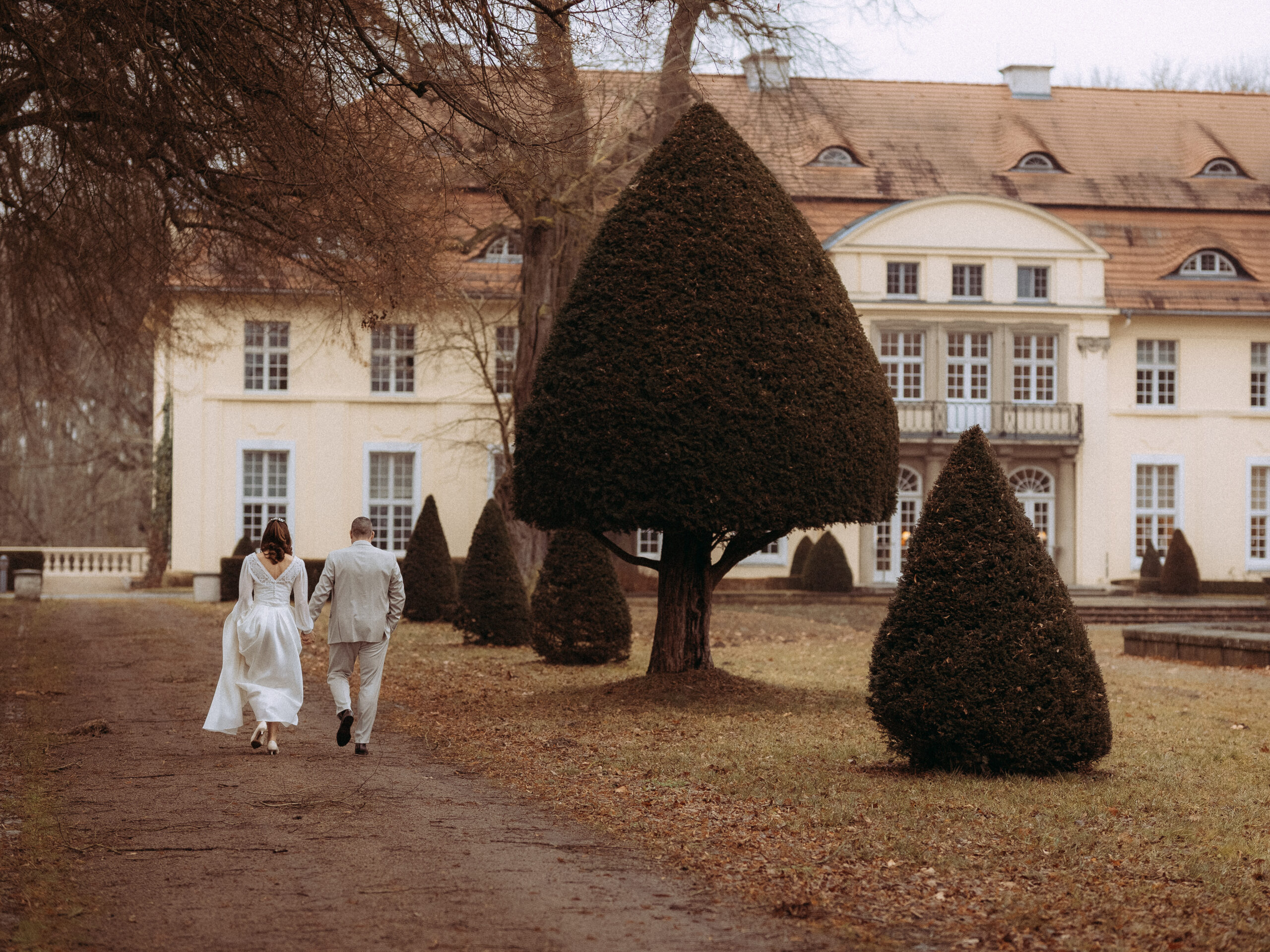 Brautpaar spaziert vor Schloss mit Kegel-förmigen Bäumen.