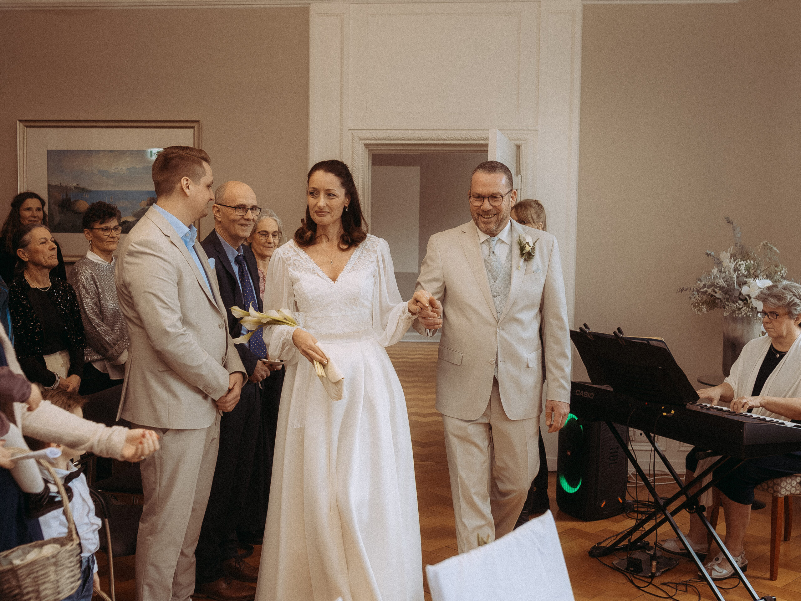 Brautpaar Hand in Hand bei Hochzeit, begleitet von Gästen und Pianistin.