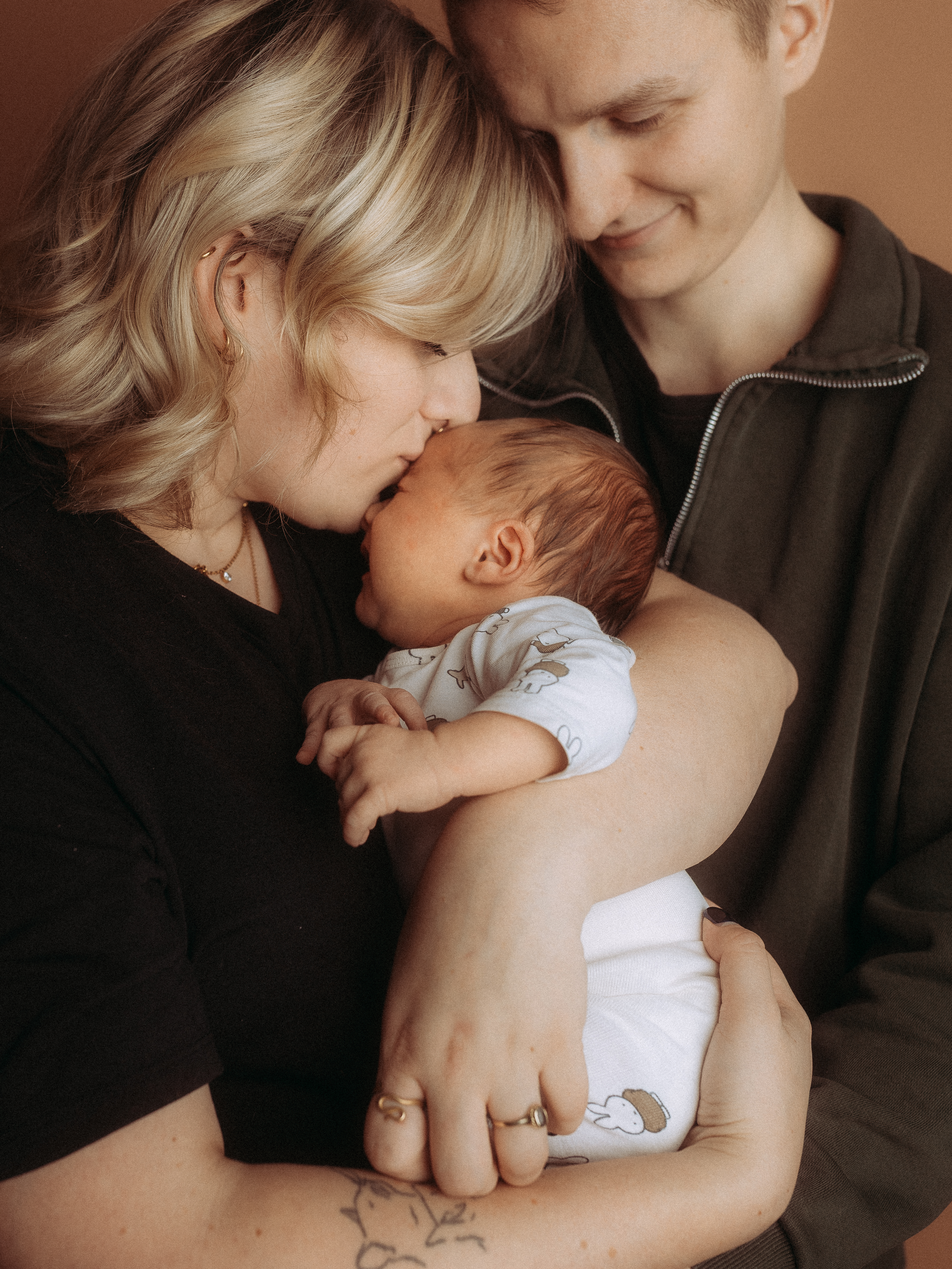 Liebevolle Familie: Mutter küsst Baby, Vater schaut zu. Glückliche Eltern mit Neugeborenem.