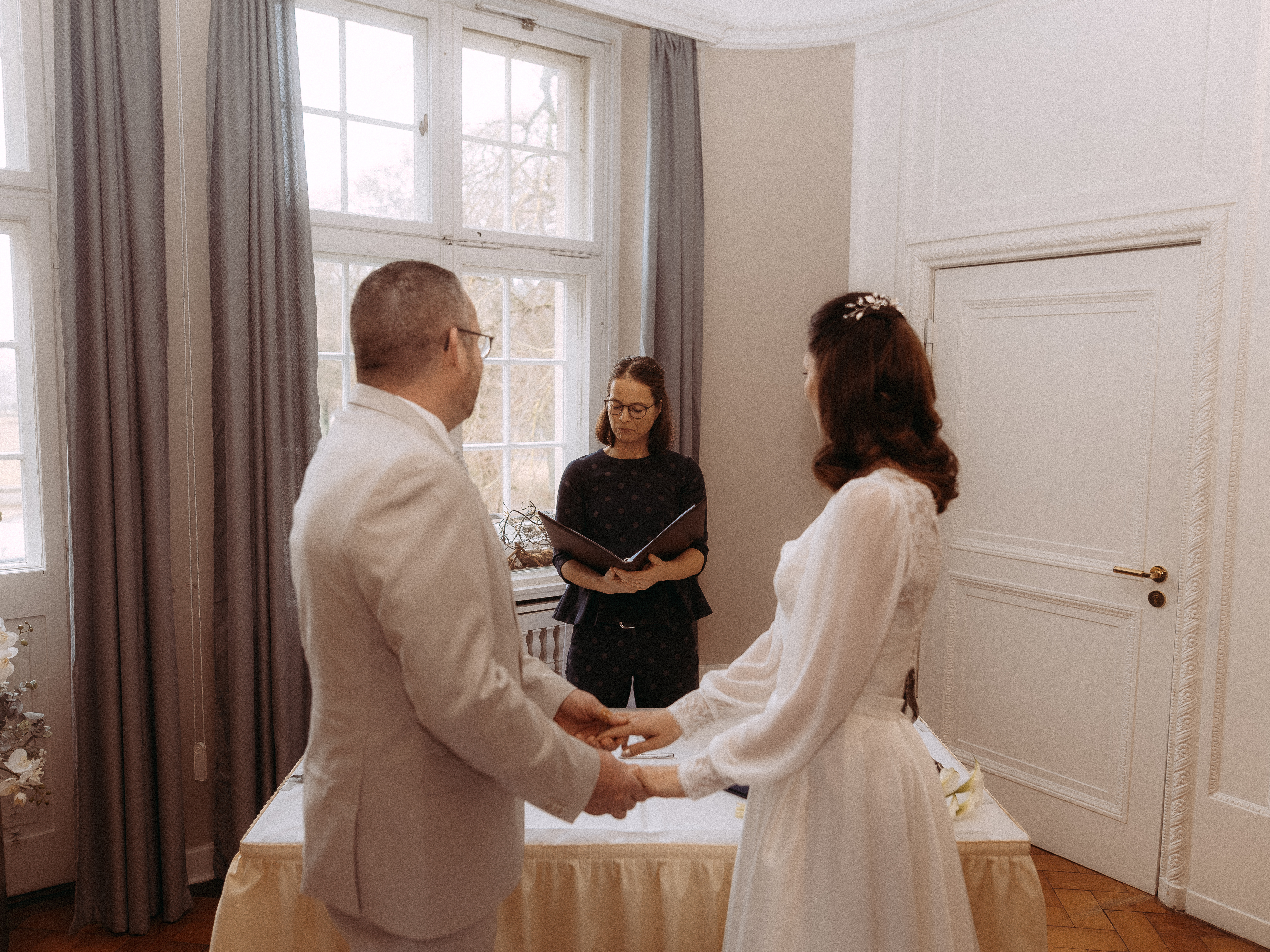 Brautpaar hält Hände bei intimer Trauung mit Standesbeamtin im Hintergrund.