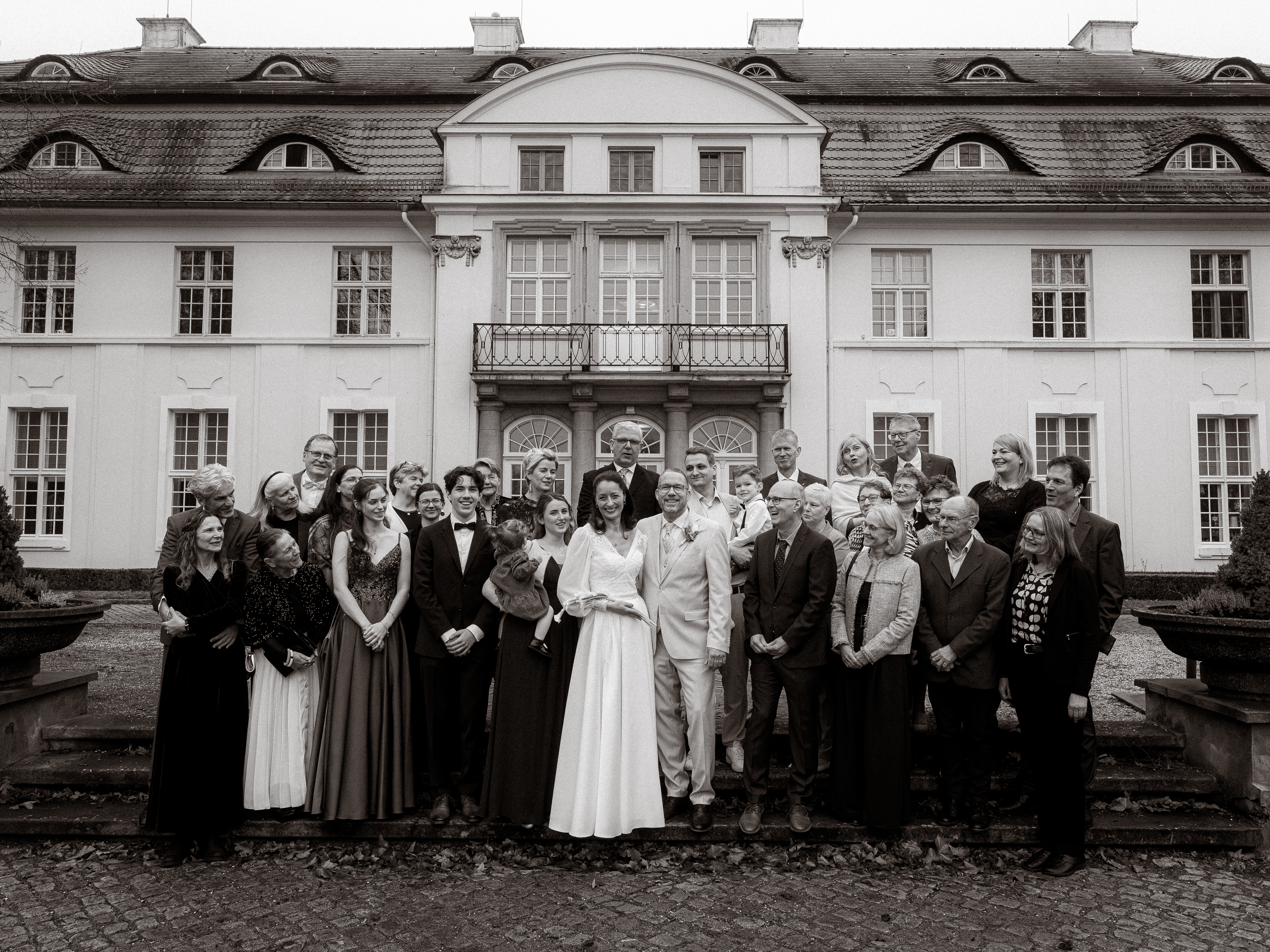 Hochzeitsgesellschaft vor einem Schloss. Brautpaar im Mittelpunkt, festliche Kleidung, klassische Architektur im Hintergrund.