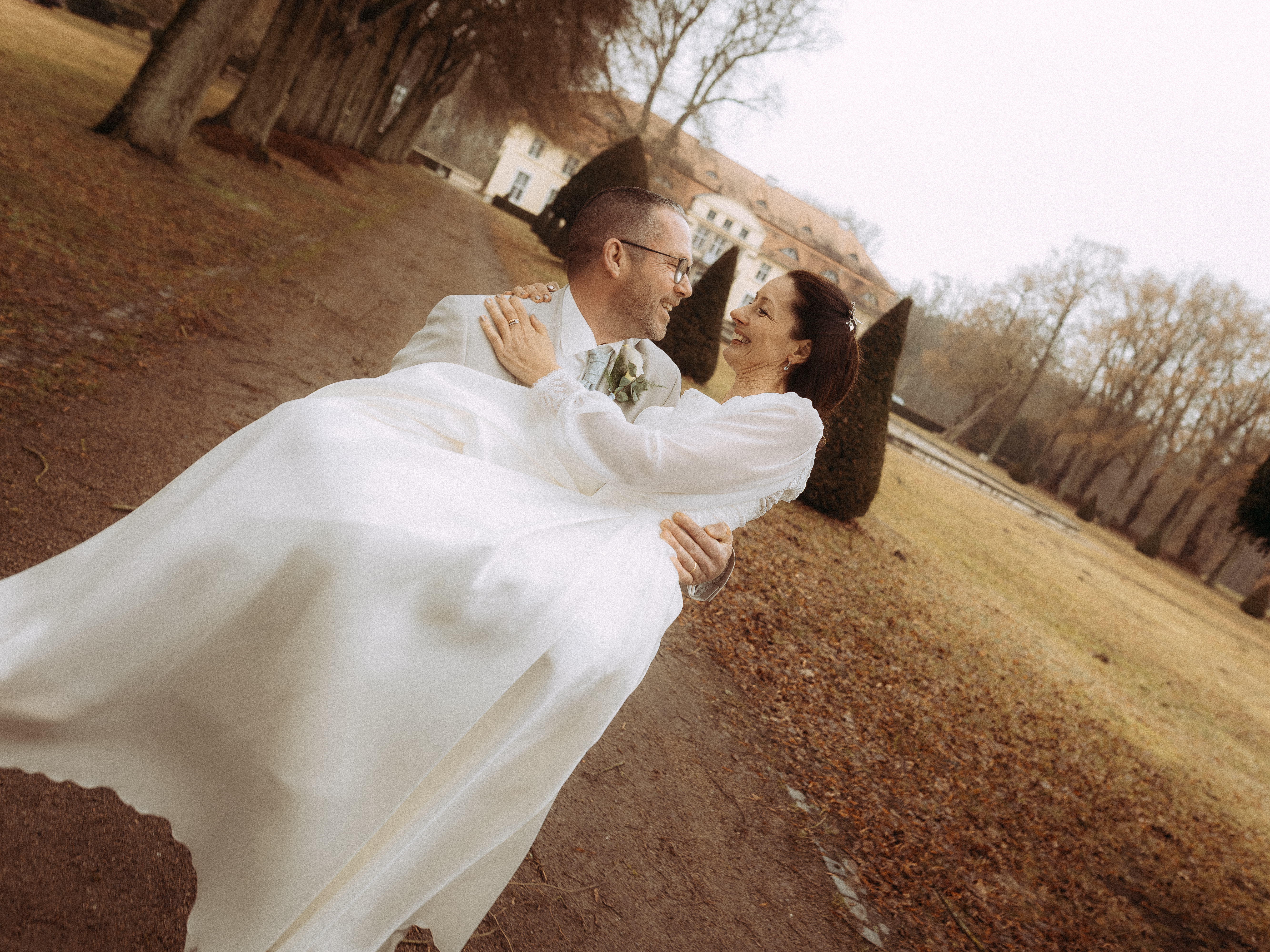 Ein glückliches Paar in Hochzeitskleidung lacht in einer sonnigen Parklandschaft