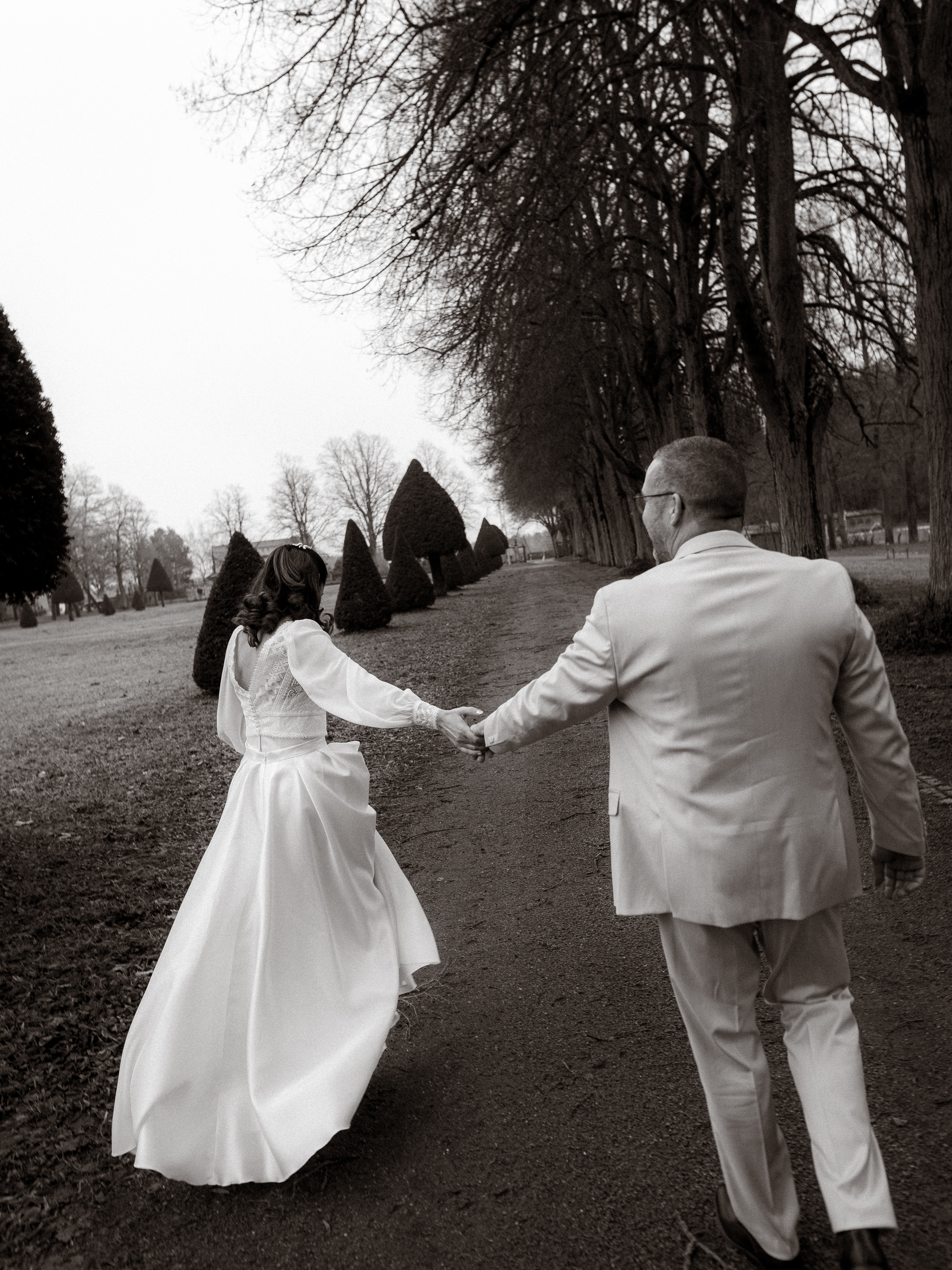 Braut und Bräutigam spazieren Hand in Hand auf einem malerischen, von Bäumen gesäumten Weg an ihrem Hochzeitstag