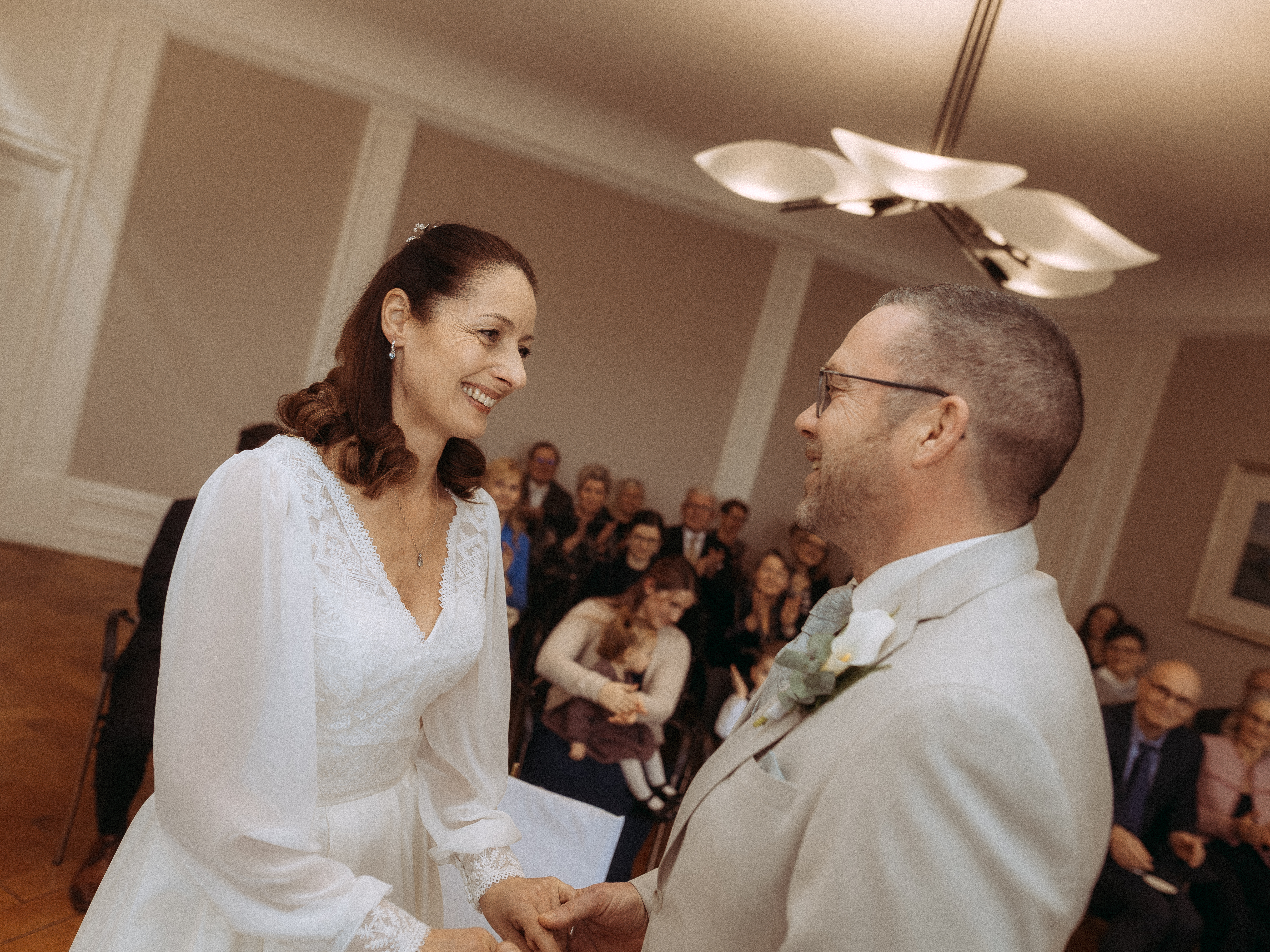 Brautpaar hält Hände bei Hochzeit. Glückliche Frau im weißen Kleid, Mann im Anzug. Hochzeitsgäste im Hintergrund.