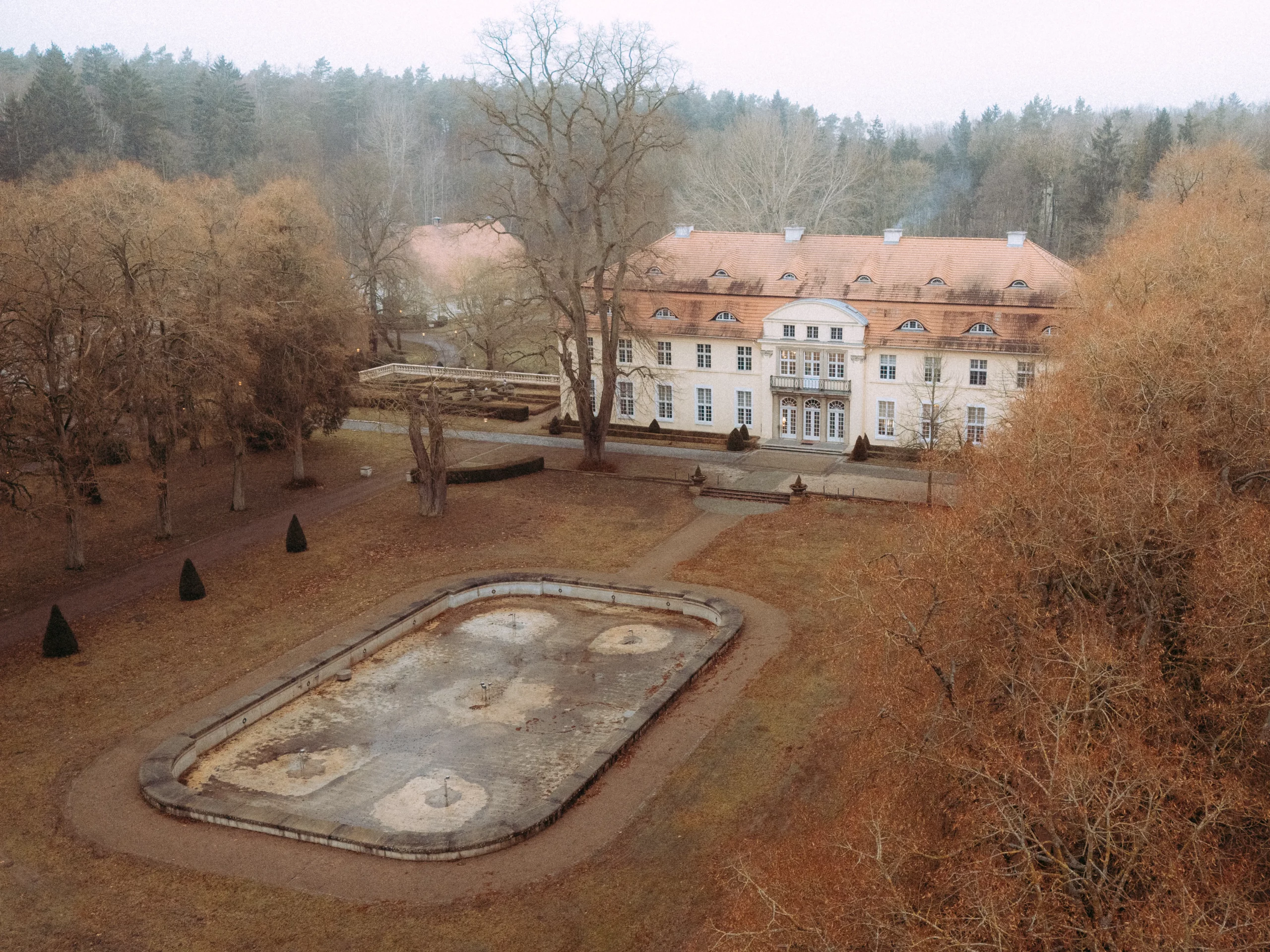 Ein großes, elegantes Herrenhaus mit rotem Dach steht hinter einem blattlosen, herbstlichen Garten. Hochzeitsfotograf MV kann hier atemberaubende Momente einfangen, mit einem leeren rechteckigen Brunnenbecken, umgeben von braunen Bäumen und Wegen.