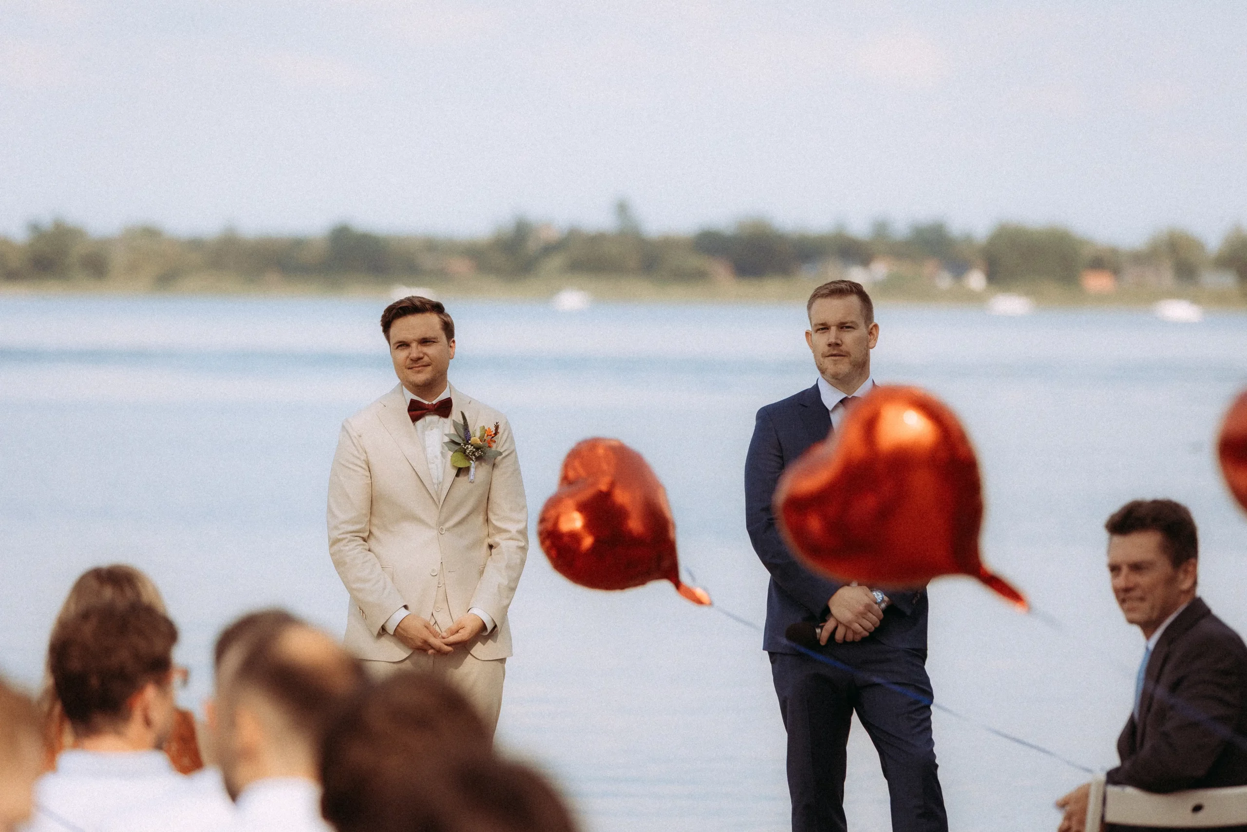 Zwei Männer stehen an einem See, einer im cremefarbenen Anzug mit Boutonniere, der andere in Marineblau. Im Vordergrund schweben rote, herzförmige Luftballons, die Gäste sind im Sitzen zu sehen - aufgenommen von einem Hochzeitsfotografen in Schwerin.
