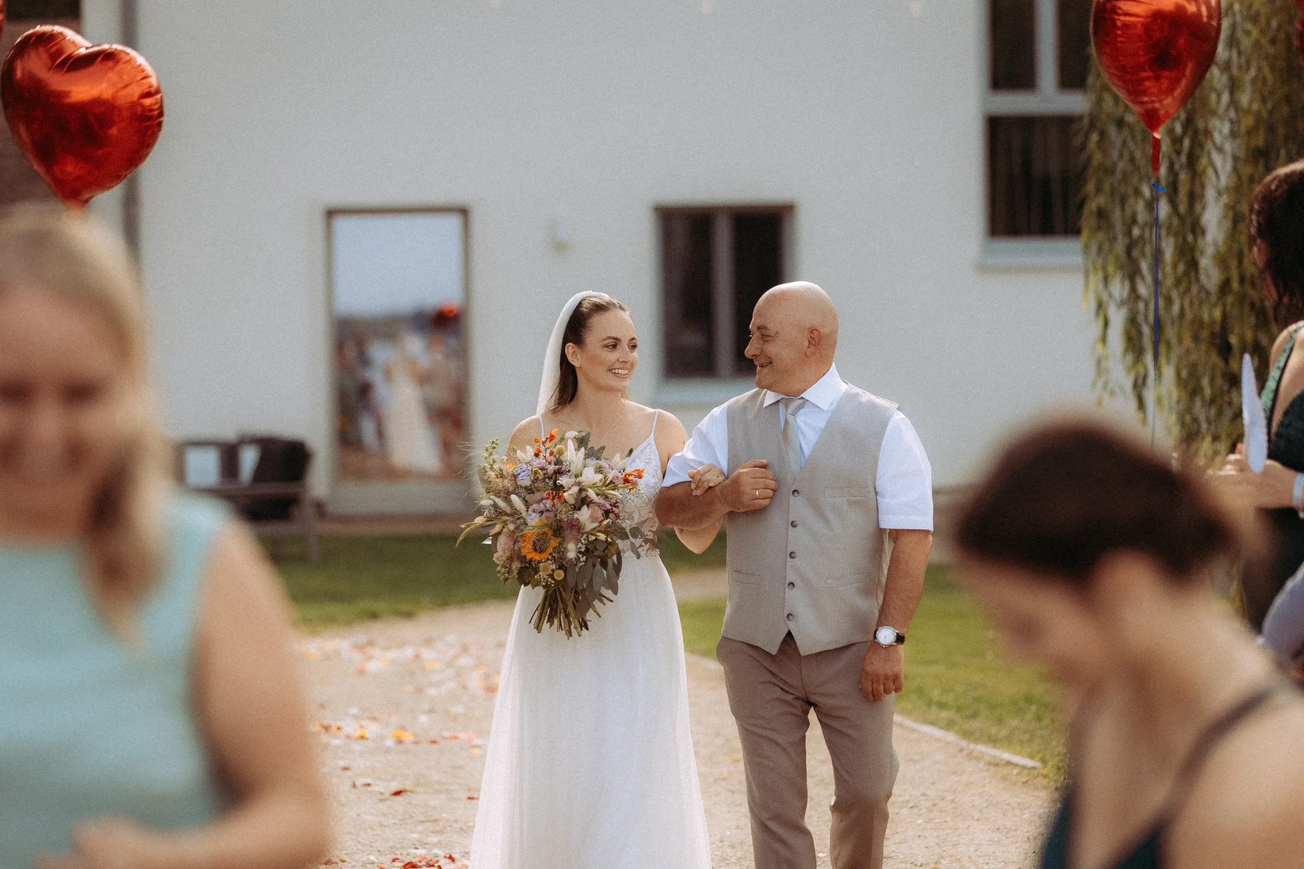 Eine Braut in einem weißen Kleid mit einem Blumenstrauß in der Hand geht Arm in Arm mit einem lächelnden Mann in einer Weste. Sie sind im Freien, umgeben von Gästen und roten herzförmigen Luftballons. Dieser Moment wurde von einem Hochzeitsfotografen in Schwerin eingefangen.