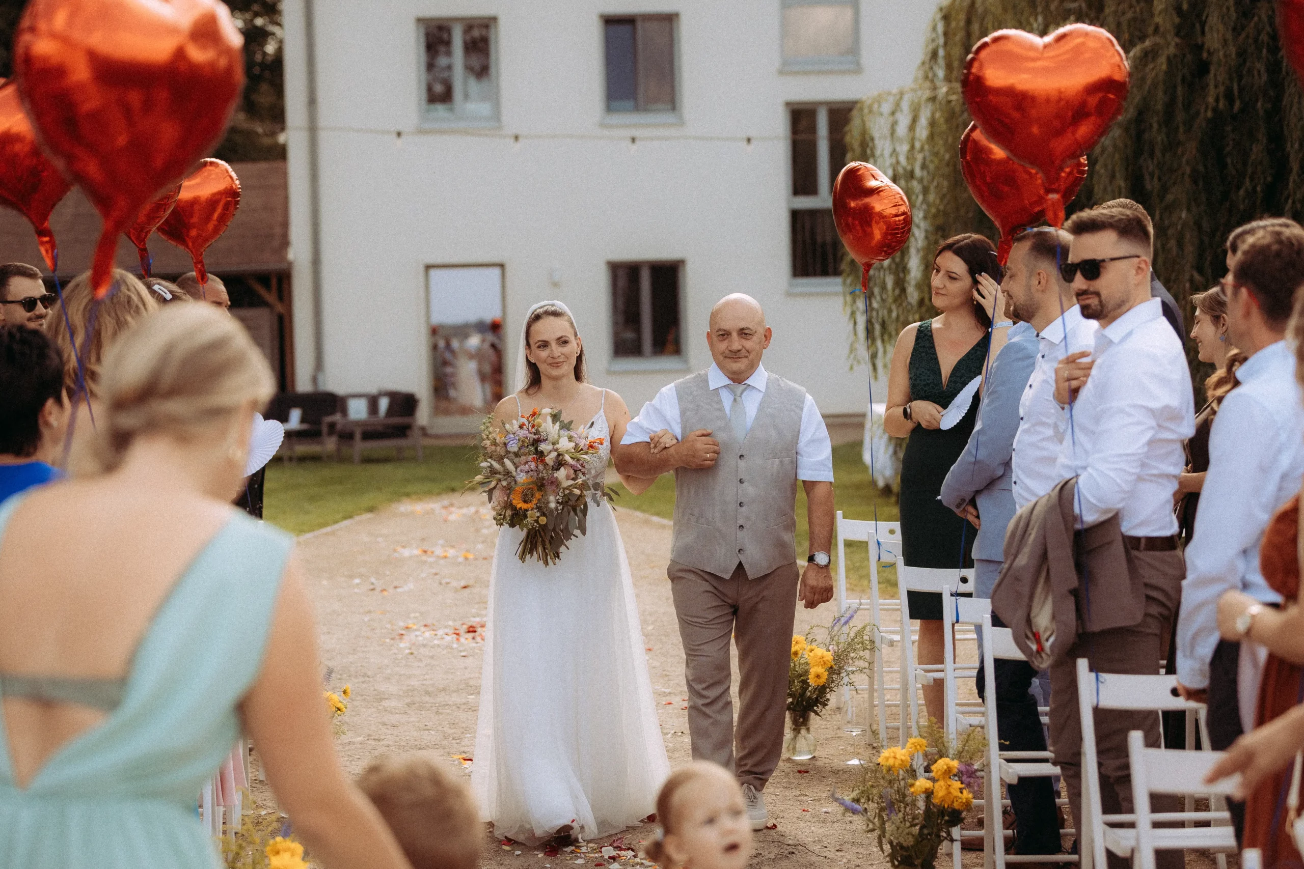 Eine Braut in einem weißen Kleid schreitet mit einem Begleiter einen Gang im Freien hinunter. Die Gäste lächeln und schauen zu, während sie rote herzförmige Luftballons halten. Die sonnige Kulisse besteht aus Blumendekorationen und einem weißen Gebäude - aufgenommen von einem Hochzeitsfotografen in Schwerin.