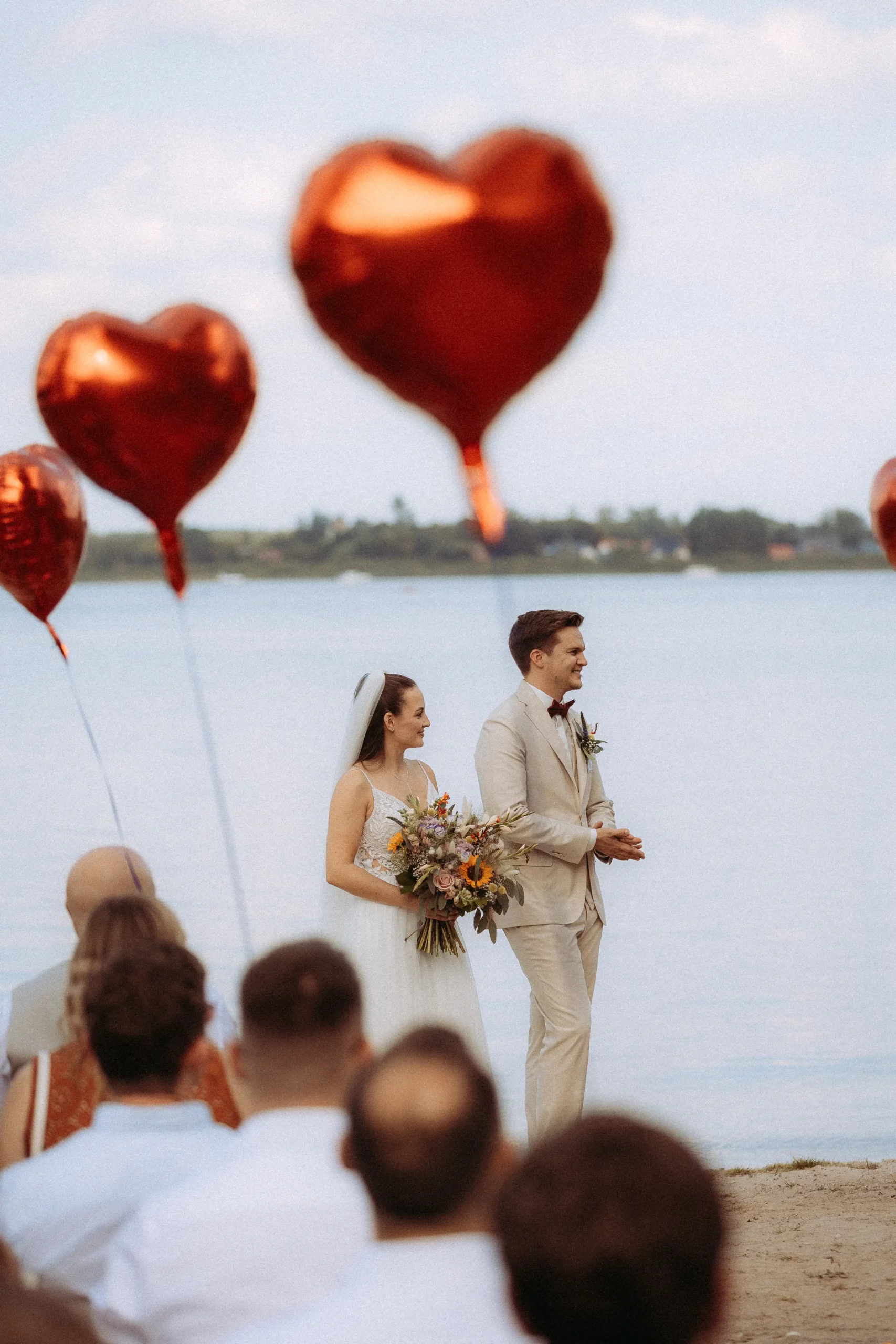 Braut und Bräutigam stehen bei einer Hochzeit im Freien zusammen am Wasser und halten Blumen, im Vordergrund sind rote herzförmige Luftballons zu sehen, dahinter sitzen die Gäste. Aufgenommen von einem Hochzeitsfotografen in Schwerin.