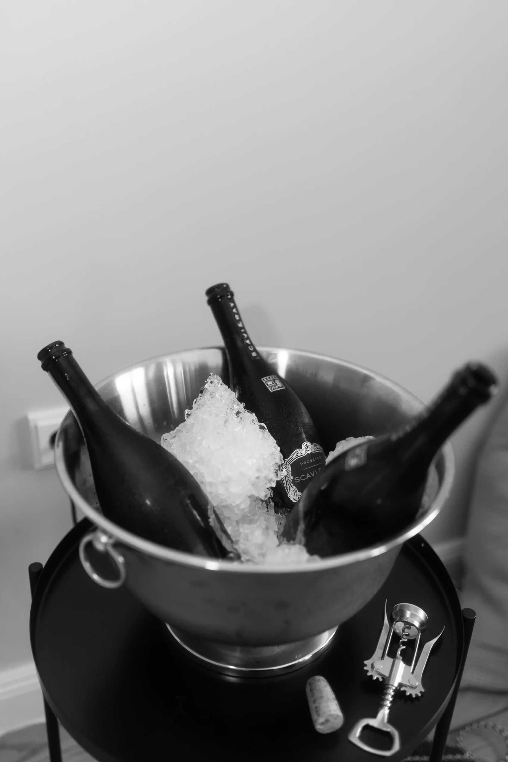 Ein mit Eis gefüllter Metalleimer und drei Weinflaschen stehen auf einem kleinen Tisch. Ein Korken, ein Korkenzieher und ein Flaschenöffner liegen neben dem Eimer. Aufgenommen in schwarz-weiß von einem Fotografen in Schwerin.