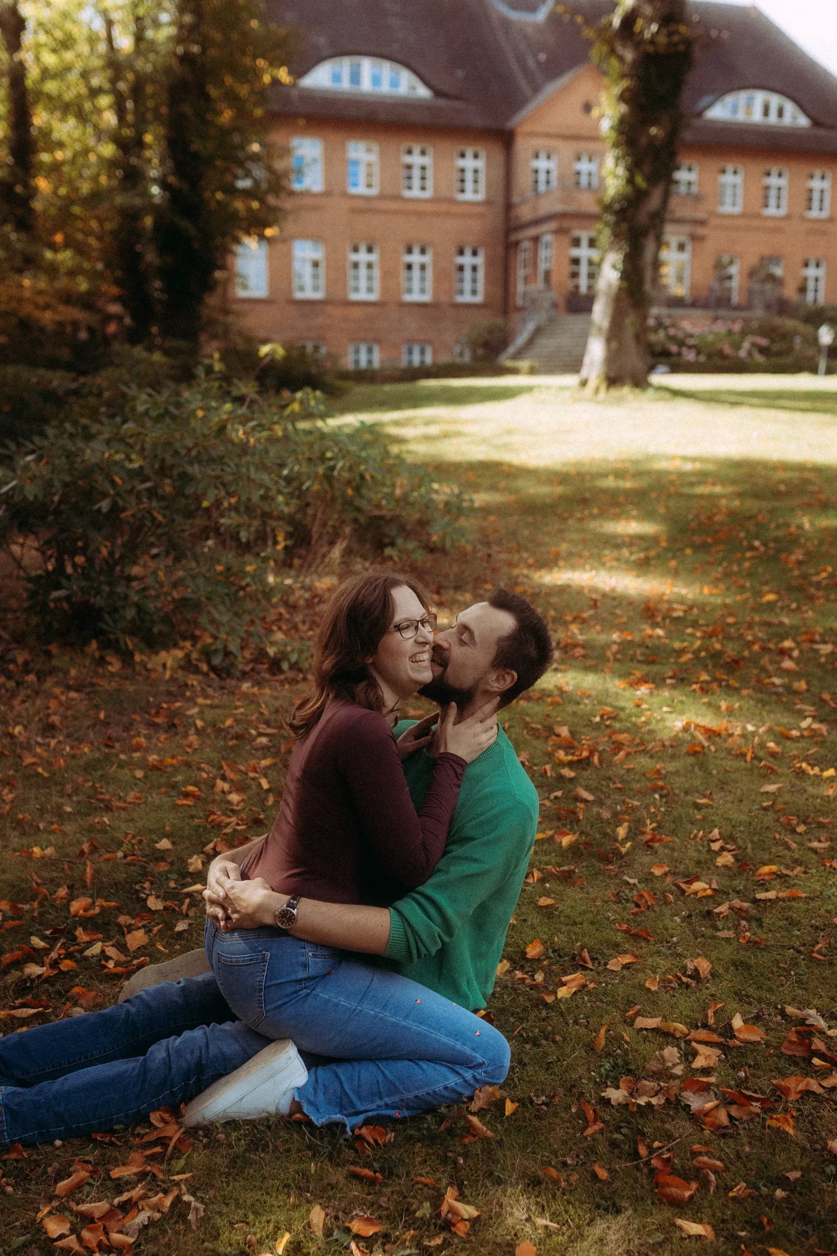Ein glückliches Paar sitzt auf einer Wiese voller Herbstlaub vor einem Backsteingebäude. Der Mann umarmt die Frau und küsst ihre Wange. Familienfotos MV.