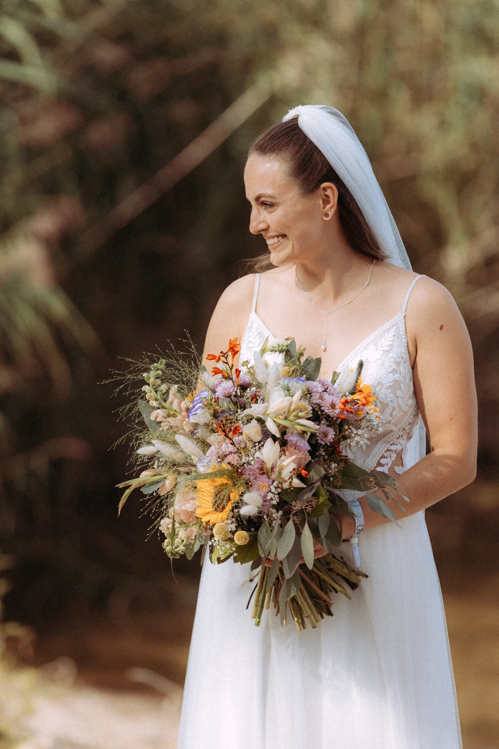 Eine lächelnde Braut in weißem Kleid und Schleier hält einen großen, bunten Blumenstrauß in der Hand, während sie im Freien im Grünen steht, aufgenommen von einem Hochzeitsfotografen in Schwerin.
