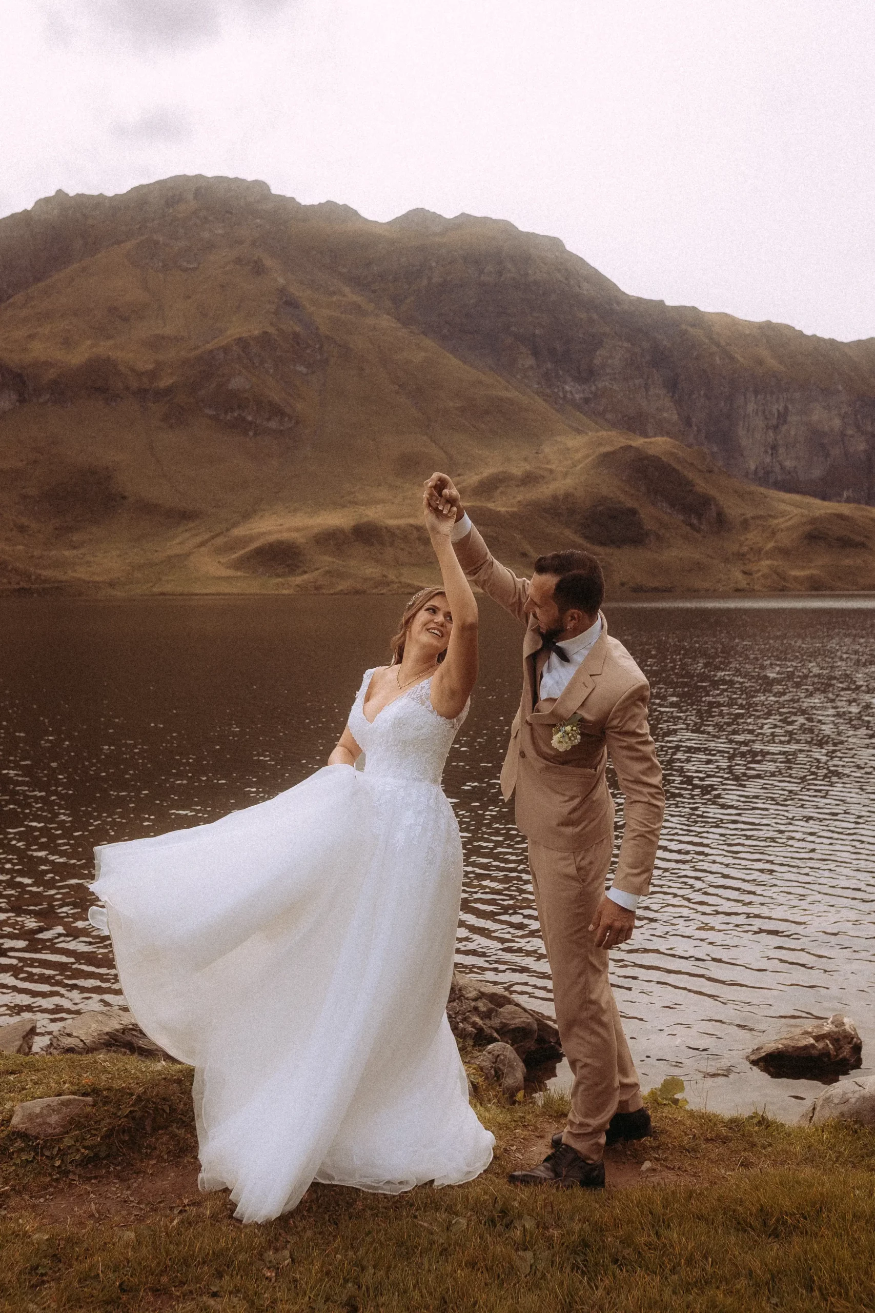 Ein strahlendes Brautpaar tanzt vor einem Bergsee. Die Braut im weißen Kleid, der Bräutigam im beigen Anzug. Hochzeitsfotograf in MV fängt die romantische Szene ein. Eine unvergessliche Hochzeit in der Natur.