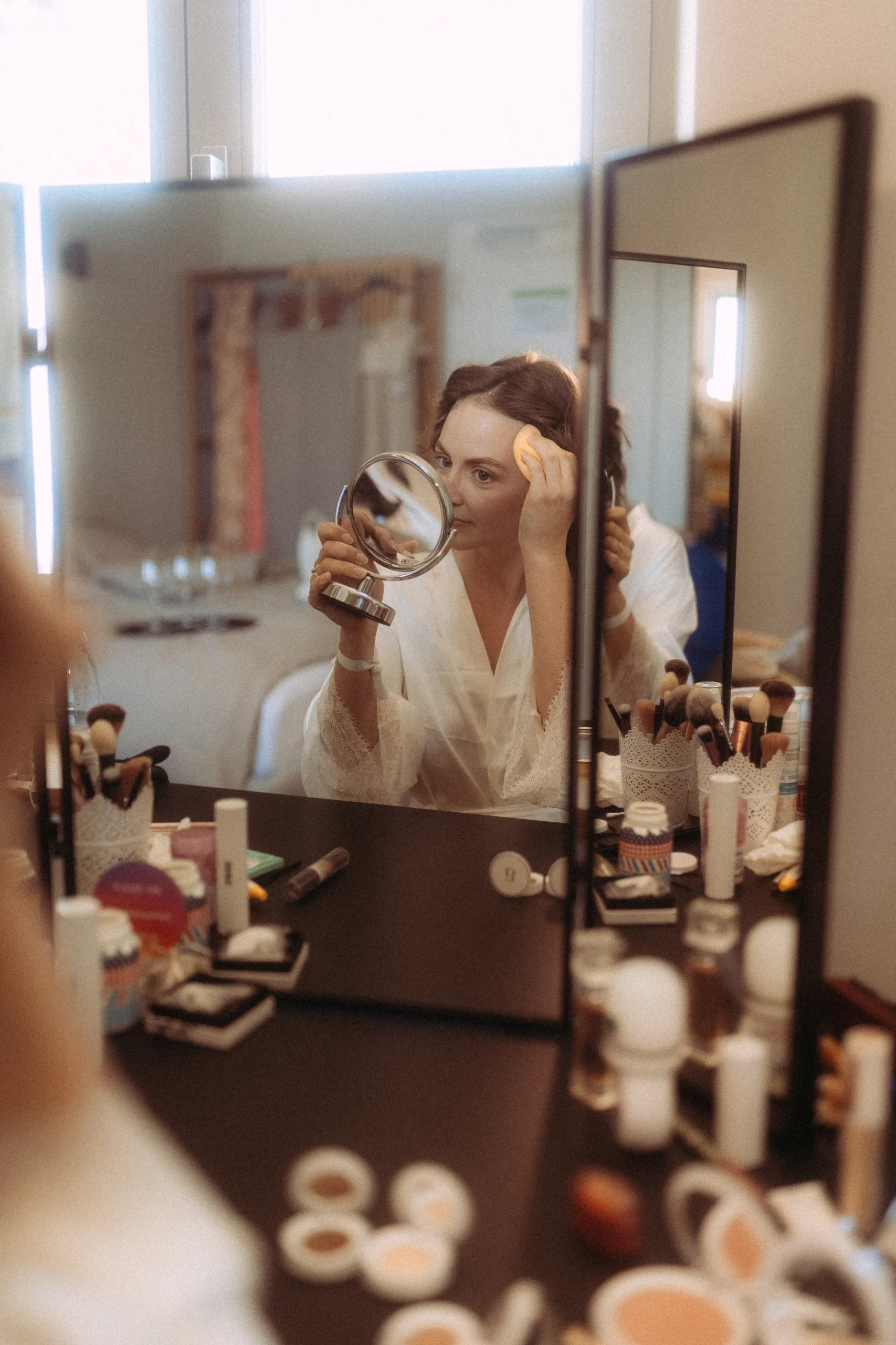 Eine Frau in weißer Robe trägt Make-up mit einem runden Schwamm auf und blickt in einen Handspiegel an ihrem Waschtisch, umgeben von Make-up-Produkten und Pinseln, die sich in einem großen dreifach gefalteten Spiegel spiegeln - aufgenommen von ein Fotograf in Schwerin.
