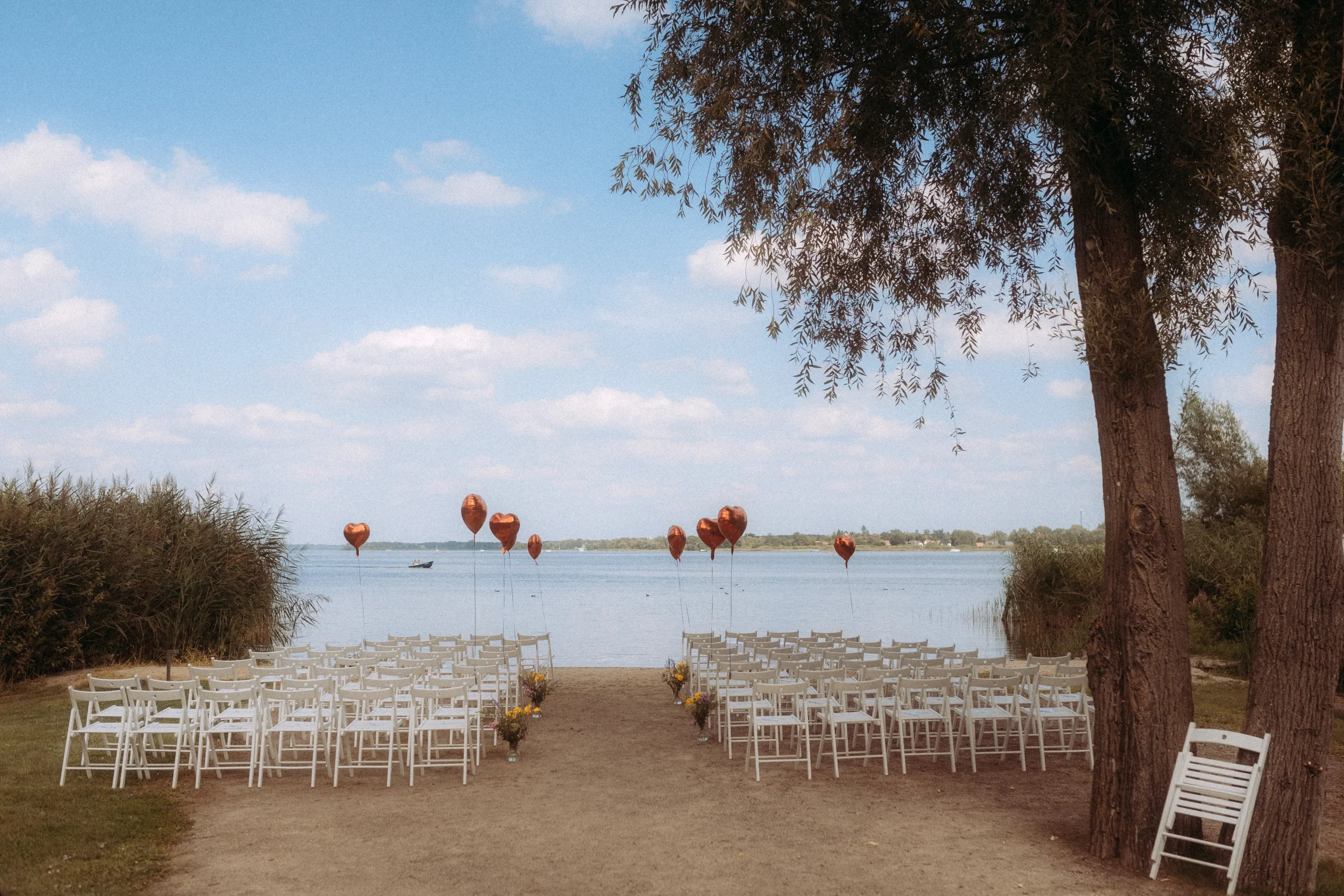 Romantische Hochzeit am See in MV. Weiße Stühle sind für die Zeremonie aufgestellt, dekoriert mit Blumen und orangefarbenen Ballons. Hochzeitsfotograf in MV hielt diesen Moment fest.