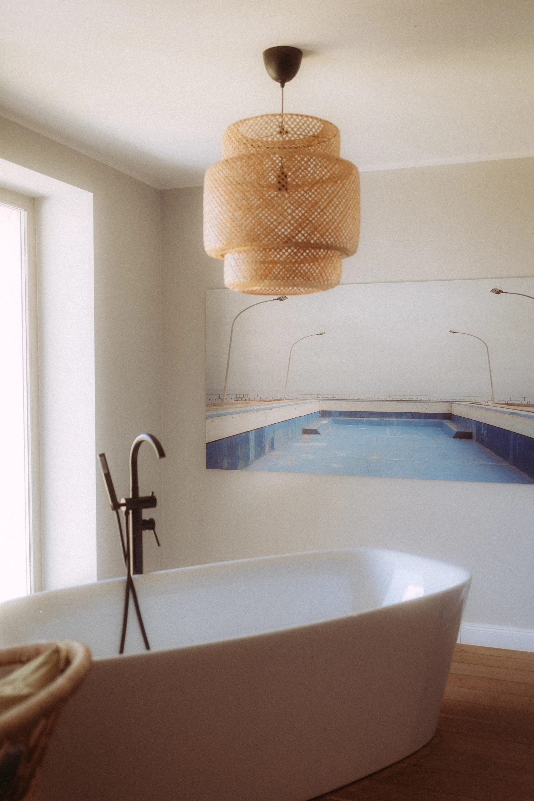 Ein modernes Badezimmer mit einer weißen freistehenden Wanne, einer schwarzen Bodenarmatur, einer Pendelleuchte aus geflochtenem Rattan und einem großen Wandbild eines Schweriner Fotografen, das ein leeres Schwimmbad mit Straßenlaternen zeigt. Natürliches Licht fällt von links ein.